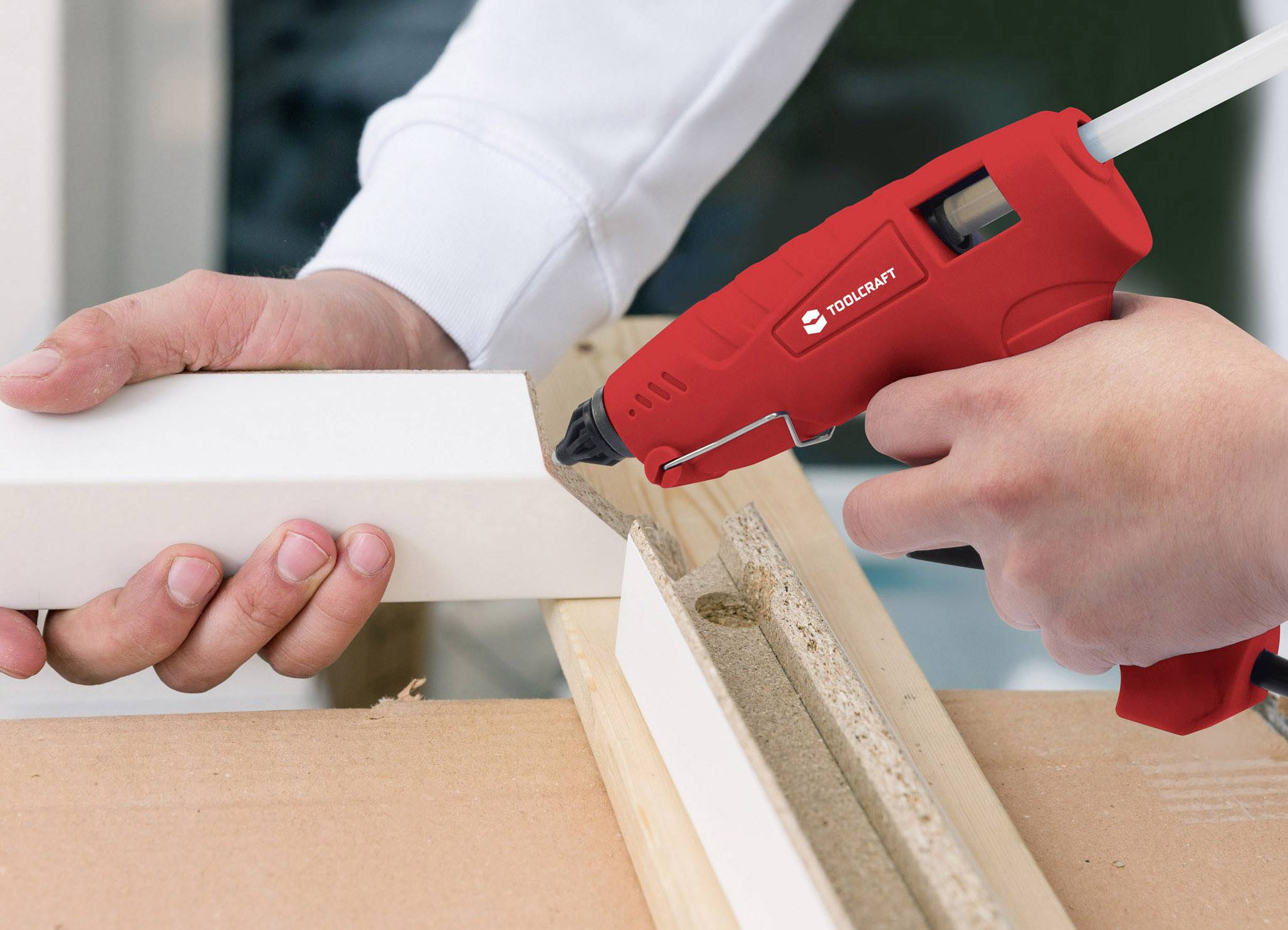 A person is using a red hot glue gun to connect two white pieces of wood. Hands are holding the materials in position.