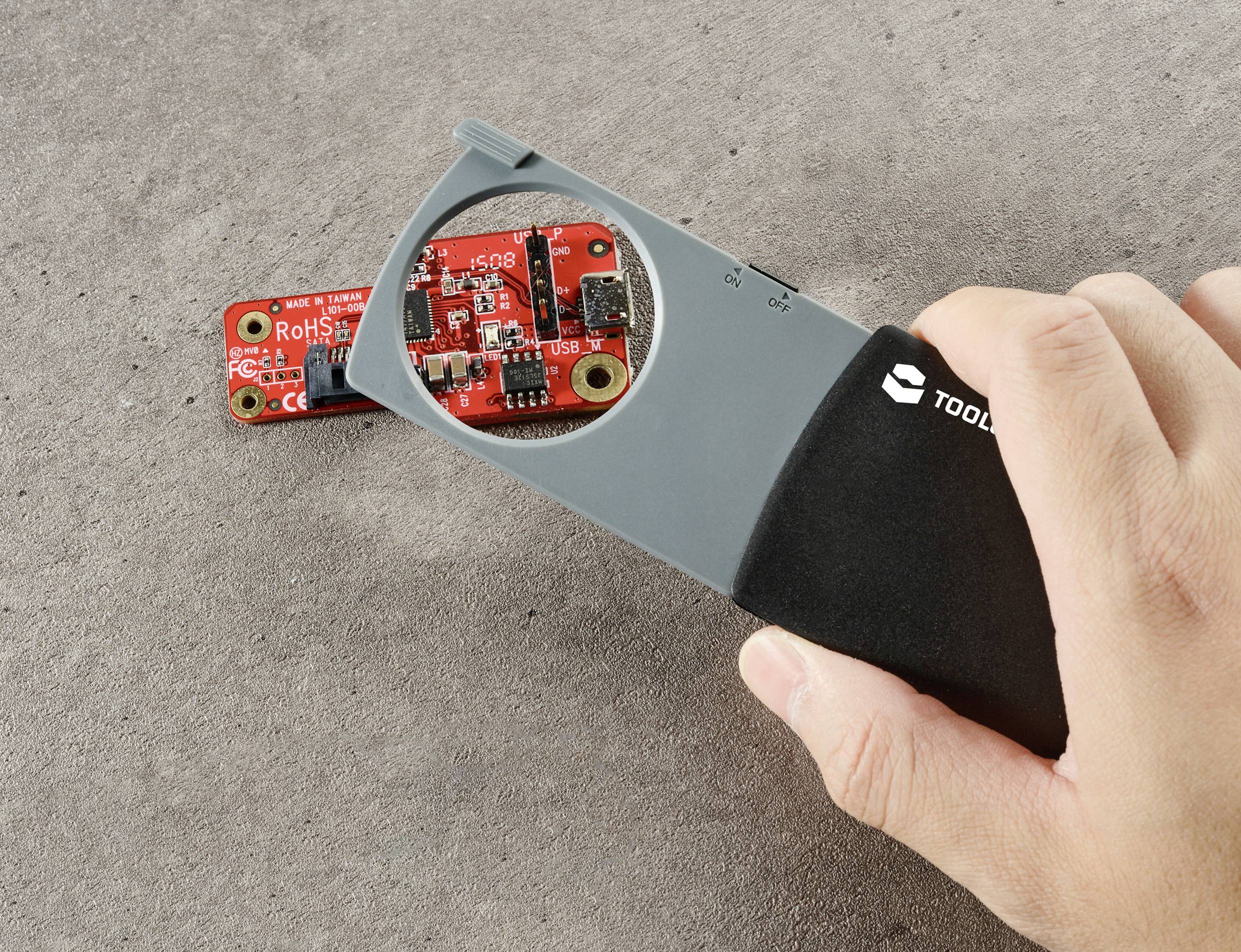 A hand holds a magnifying glass over a small red circuit board, with electronic components and inscriptions visible.