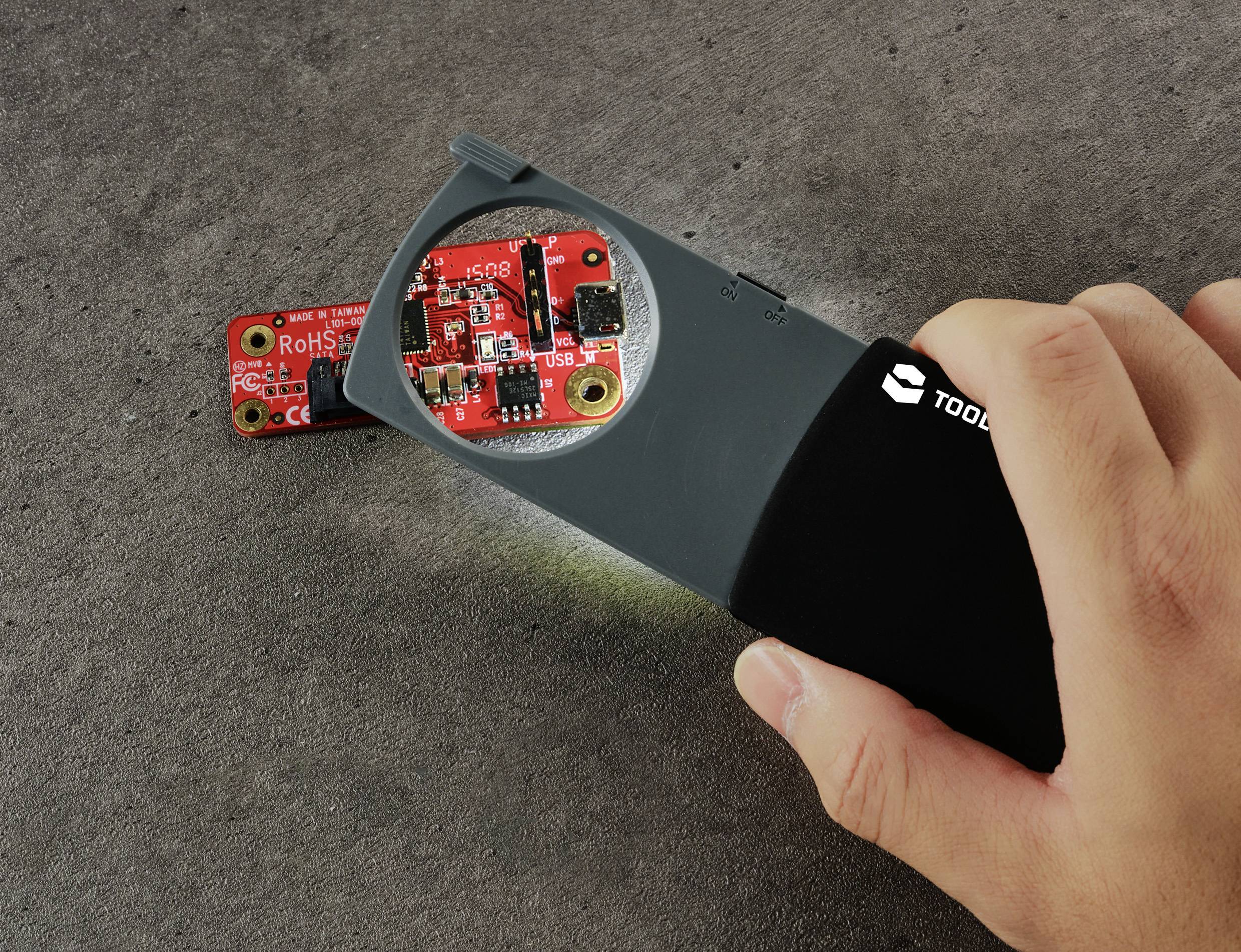 A hand holds a torch over a small red circuit board against a grey background, with highlighted details of the board visible.