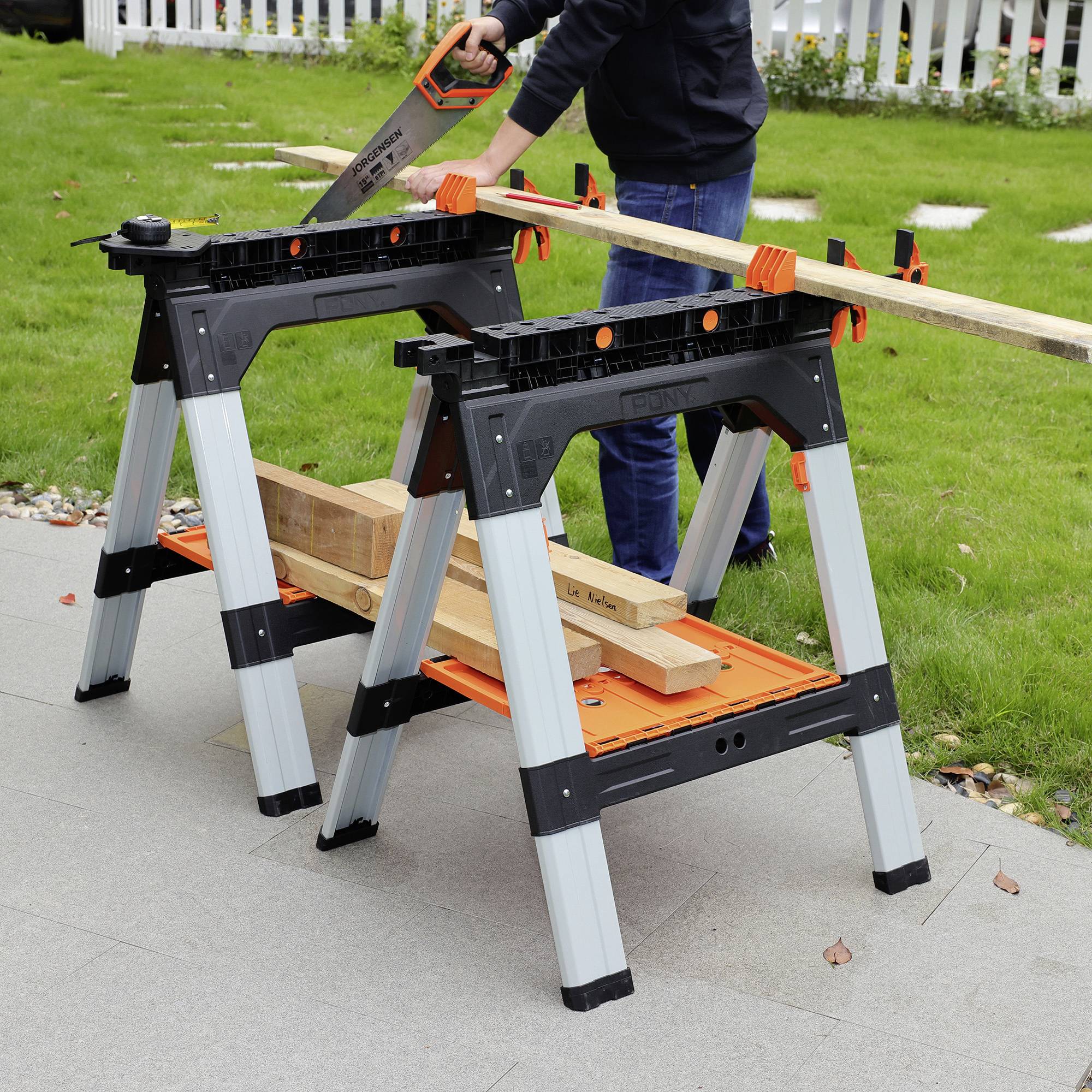 'A person is sawing a wooden batten on two sawhorses in the garden. The sawhorses are black and orange, made of plastic and metal.'