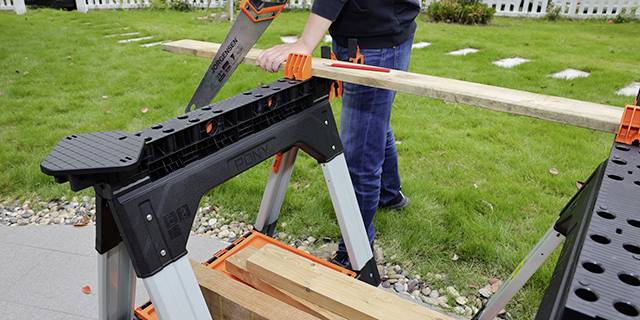 A person is sawing a wooden board on a workbench in the garden. The workbench has plastic supports, and the ground is covered with grass and paving stones.