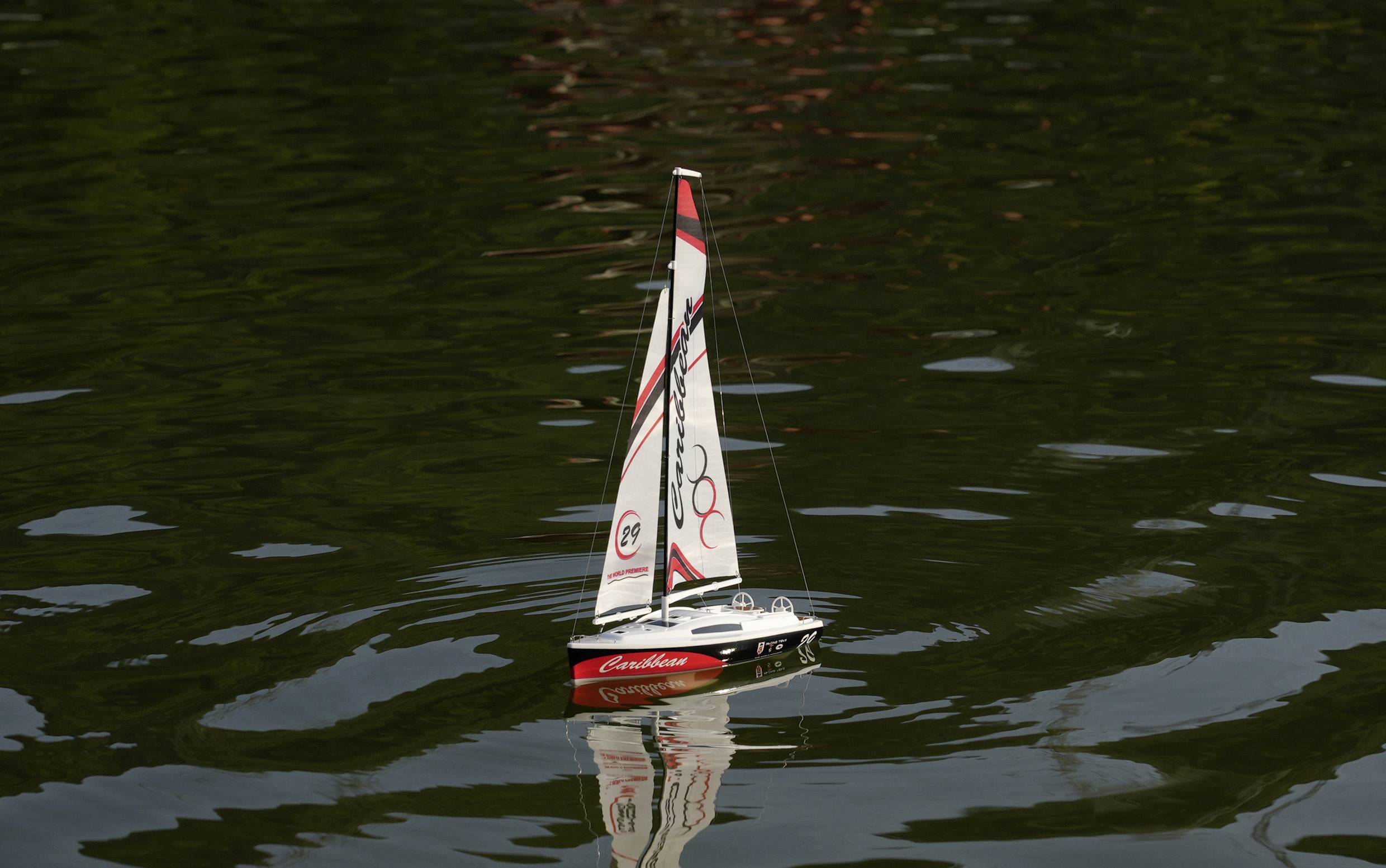 A small model sailing boat with red and white sails floats on a calm body of water.