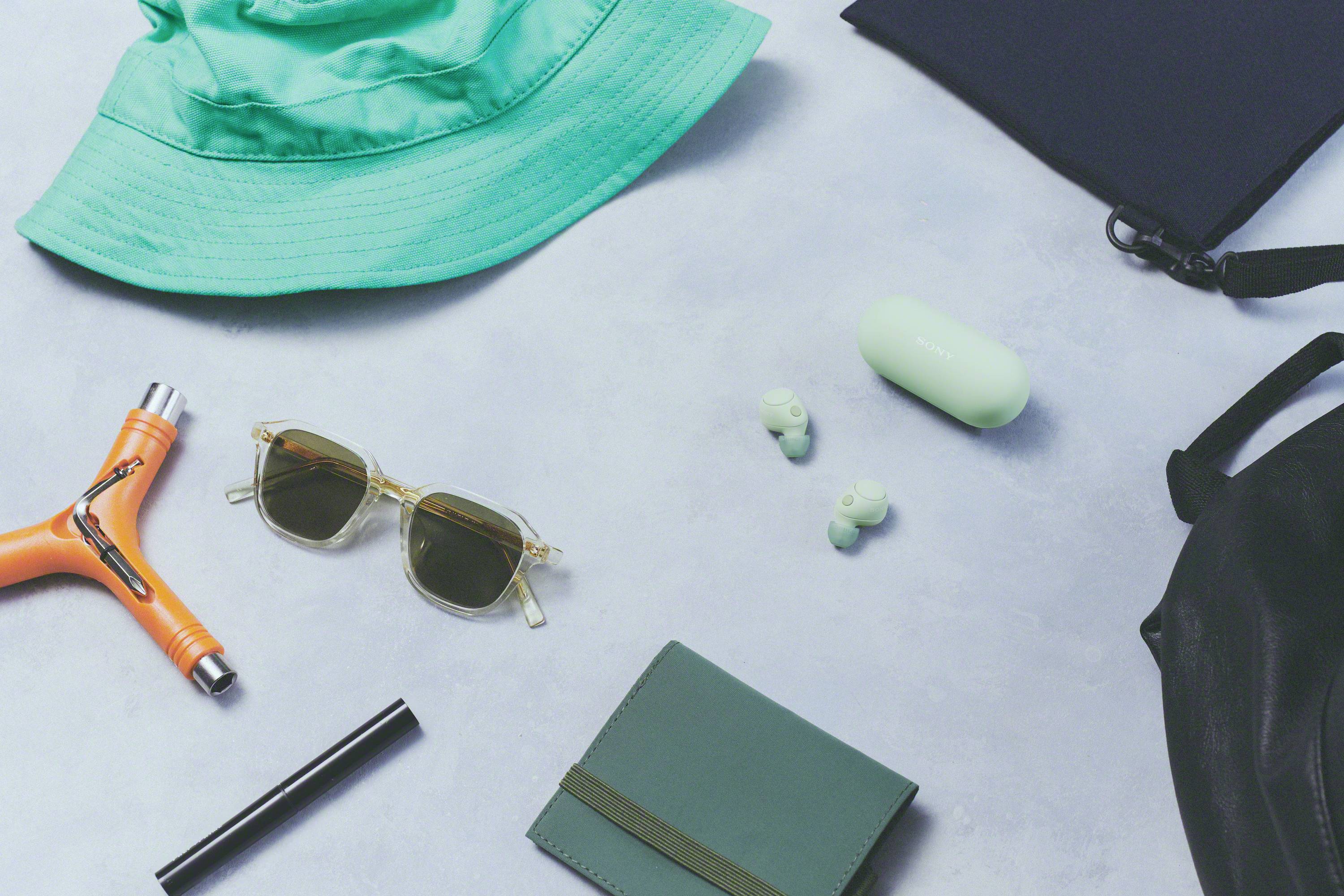 A flatlay with a green hat, sunglasses, black rucksack, orange tool, green wallet and wireless earbuds on a grey background.