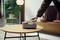 A person is picking up a round, grey-white device on a wooden table. In the background, plants and furniture can be seen in a modern room.