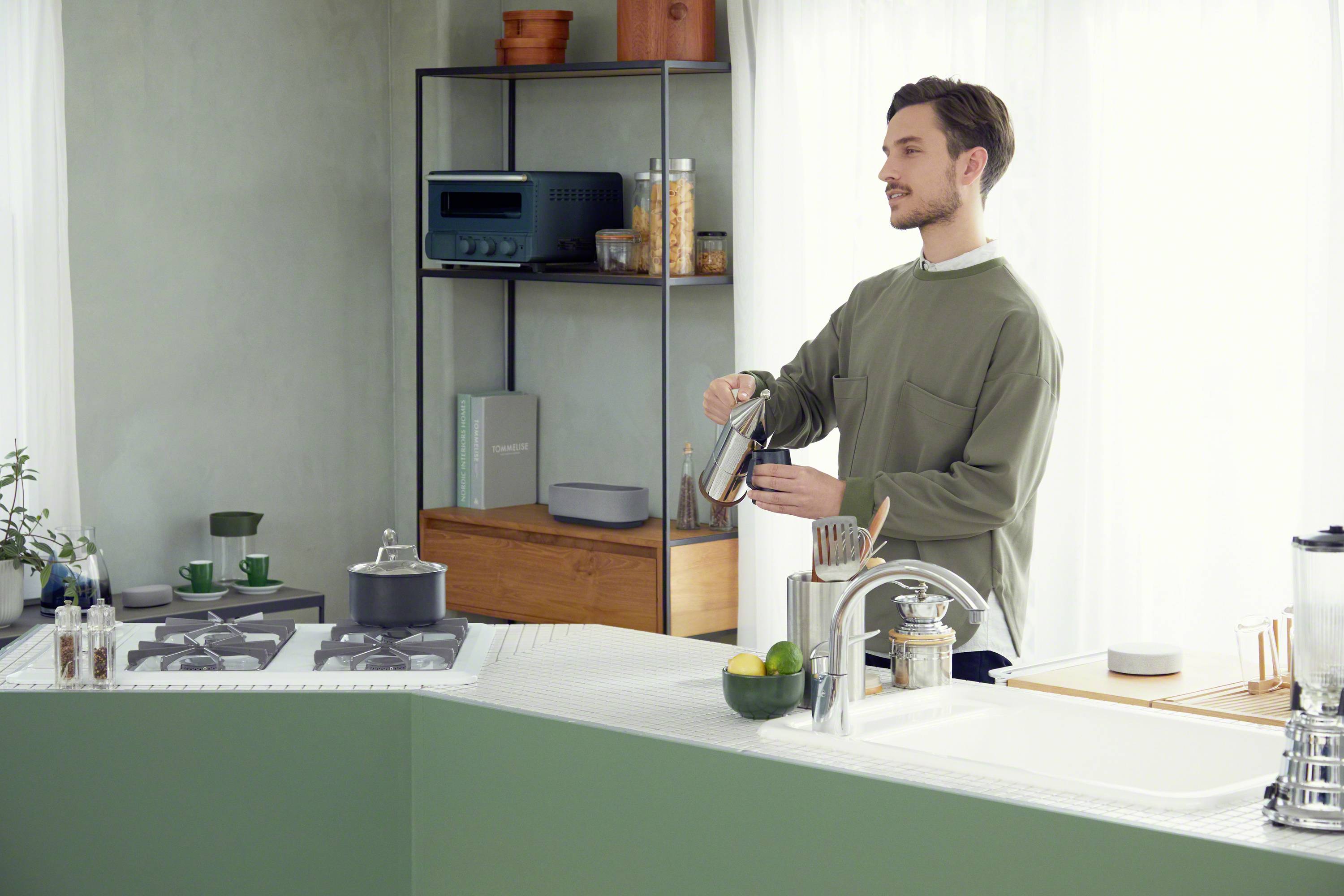 A man in a modern, bright kitchen space is pouring coffee into a cup. Kitchen utensils and shelves are visible in the background.