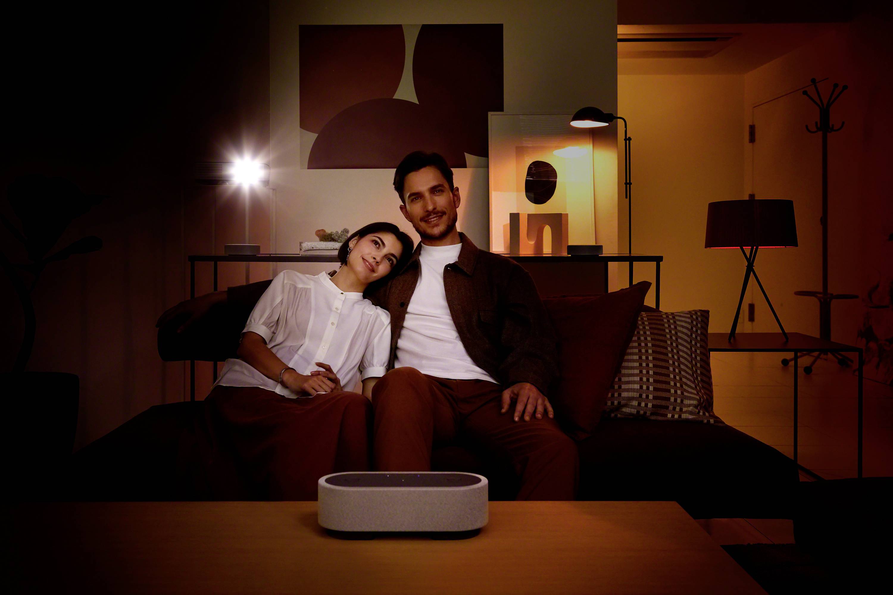 A couple sit cosily on a sofa in a room with atmospheric lighting. In front of them, a grey speaker sits on the table.