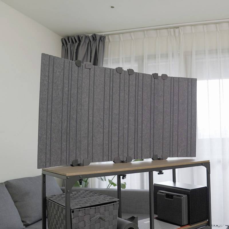 A room with a grey sofa, a window with curtains, and a table featuring an acoustic divider, plants, and a speaker.
