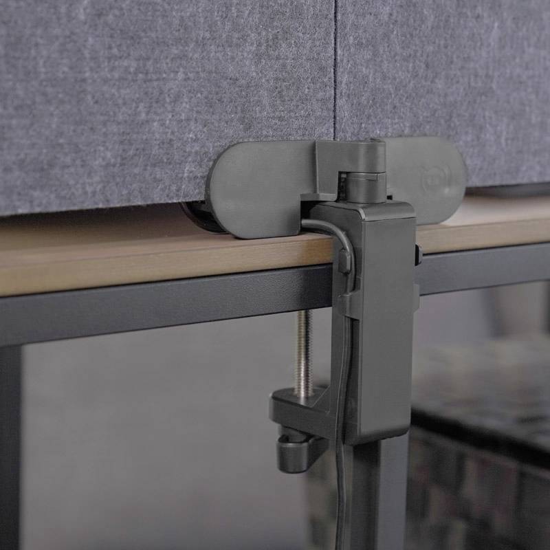 A black clamp is attached to the edge of a table to securely fix a grey partition. The surroundings are out of focus.