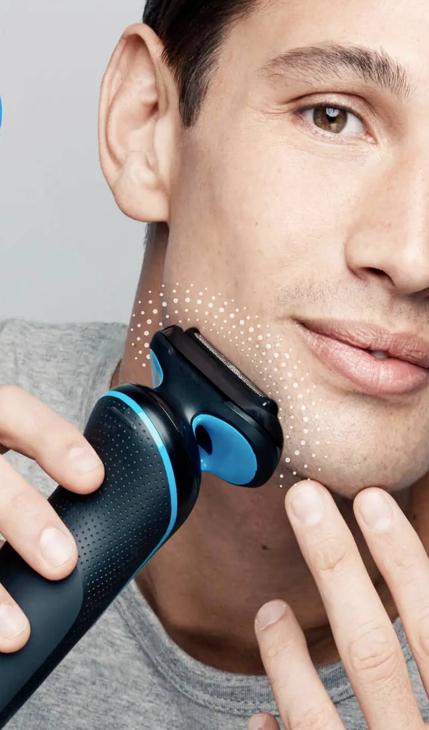 A person is shaving their face with an electric razor while smiling at the camera. It shows a close-up of the razor application.