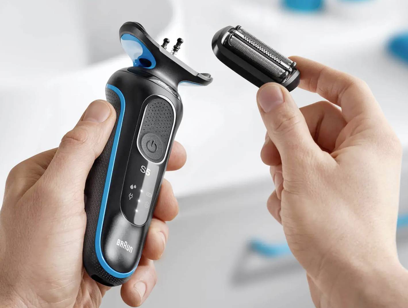 A person is holding an electric razor in their left hand and removing the shaving head with their right hand. Bathroom visible in the background.