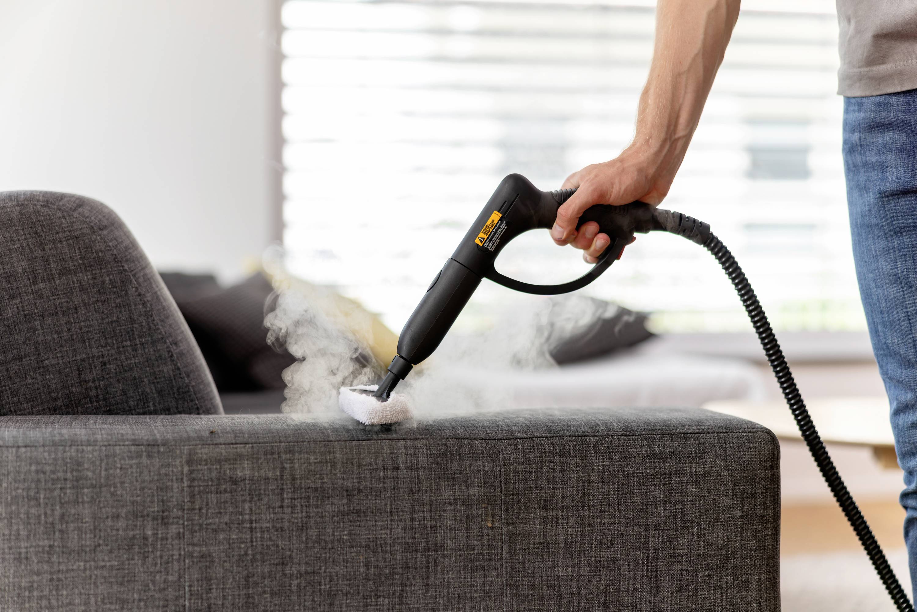 A person is cleaning a grey sofa with a steam cleaner. Steam rises from the device. Blurred room details are visible in the background.
