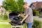 A young man is cleaning a barbecue outdoors with a pressure washer. Trees and houses are visible in the background.