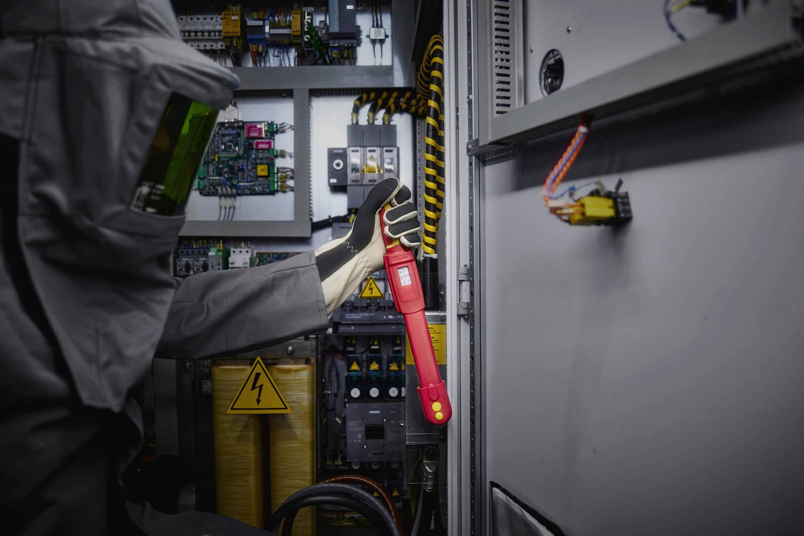 A person in protective clothing is working with an insulated tool on an open electrical cabinet. Safety signs are visible.