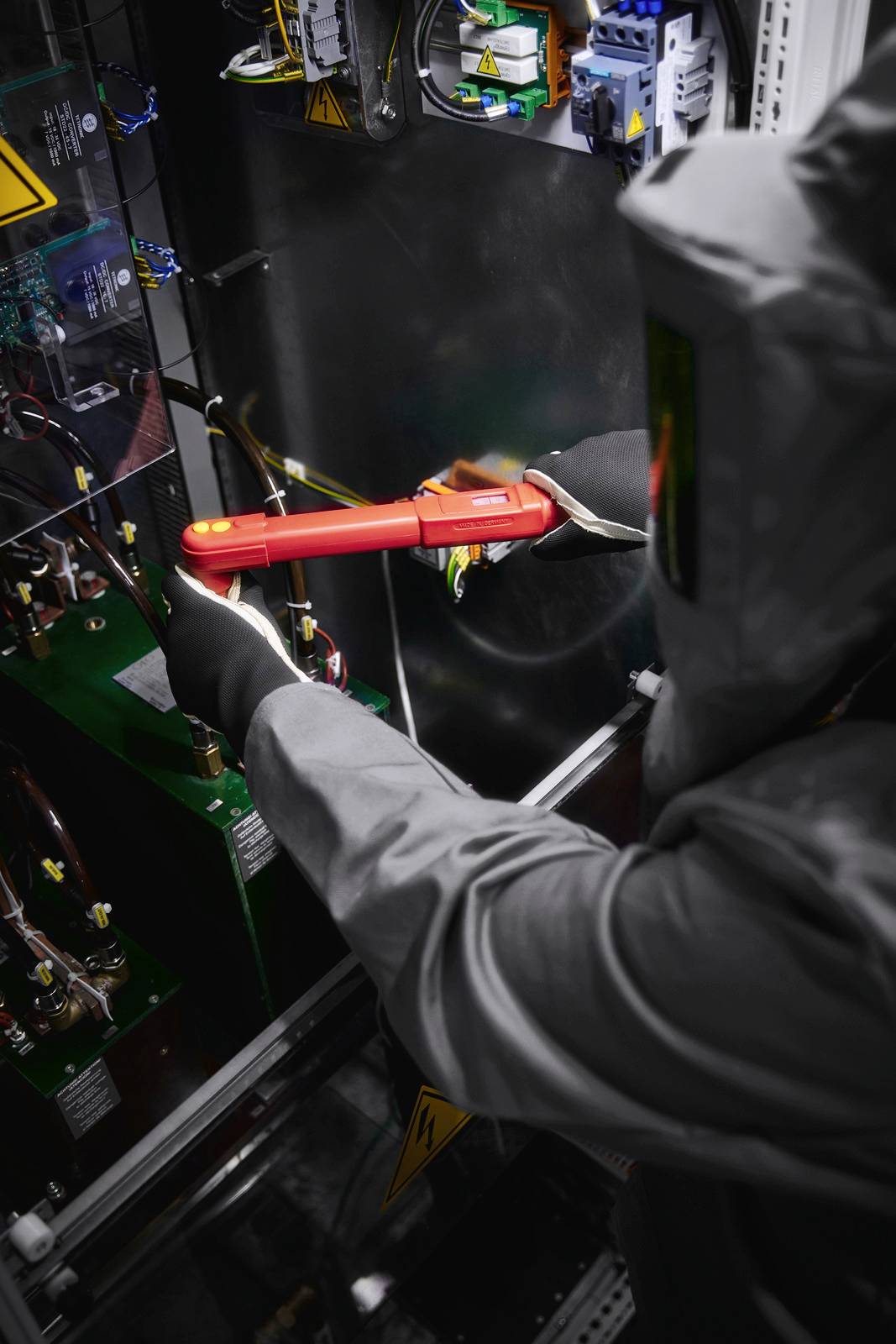 A person wearing personal protective equipment is working with a voltage tester in an electrical switchboard.