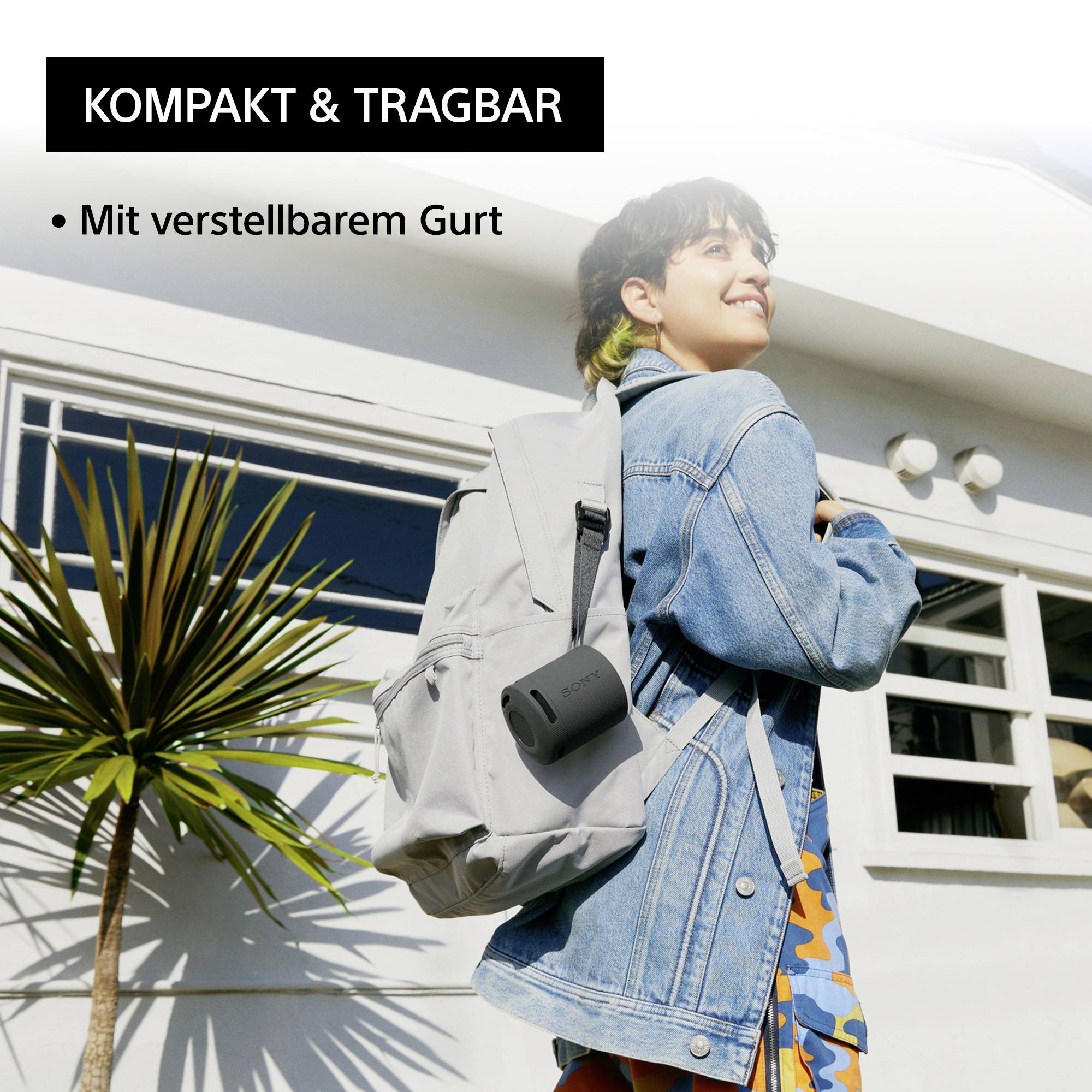 Person in a denim jacket with a grey rucksack and portable speaker smiling, standing outside next to a plant and a window. Text: 'Compact & Portable, With Adjustable Strap'.