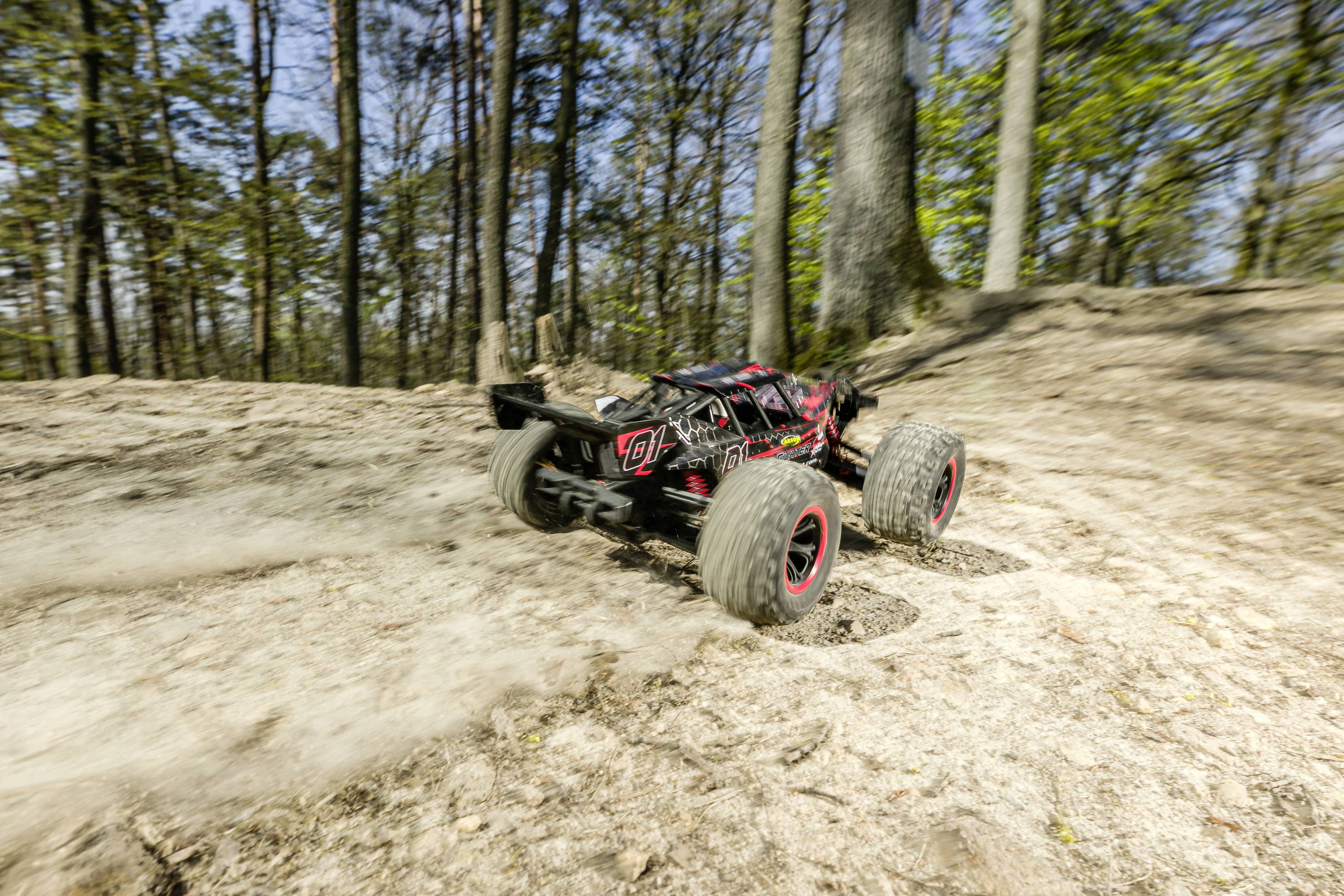 A remote-controlled car drives quickly over sandy ground in a woodland area. Trees and vegetative surroundings in the background.