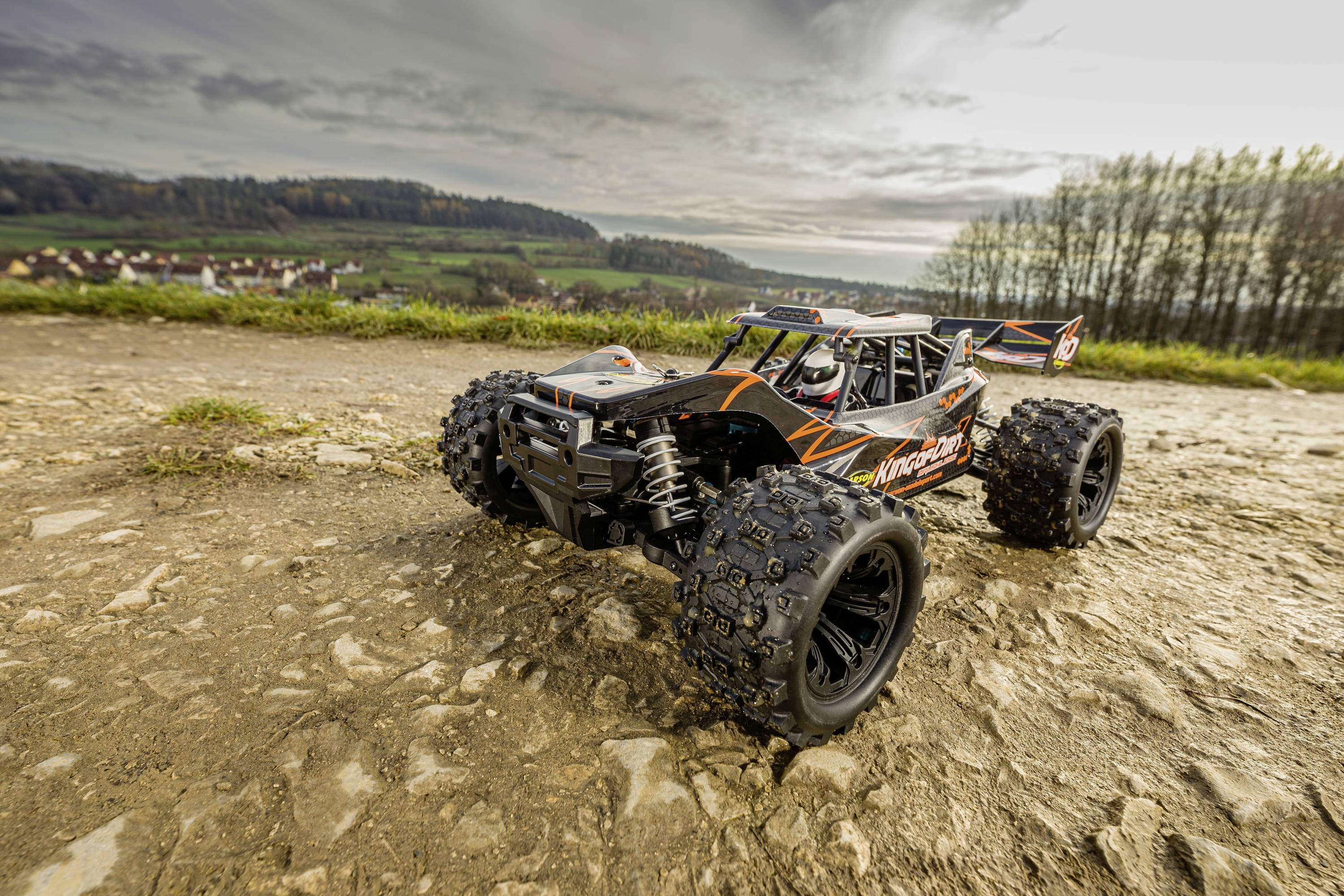 A remote-controlled car with large tyres drives along a rocky path in a rural landscape. Fields and trees can be seen in the background.