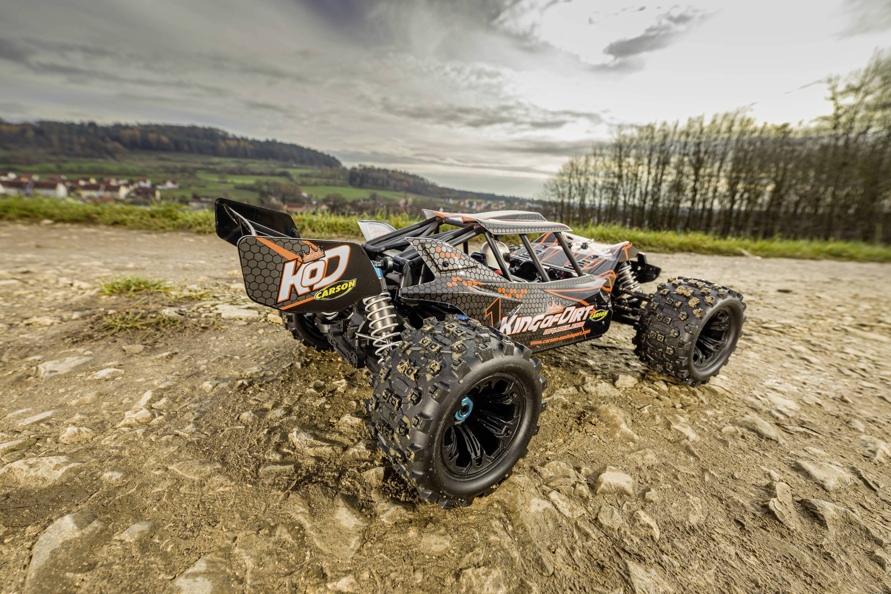 A remote-controlled car on a gravelly path outdoors, surrounded by trees and fields in the background.