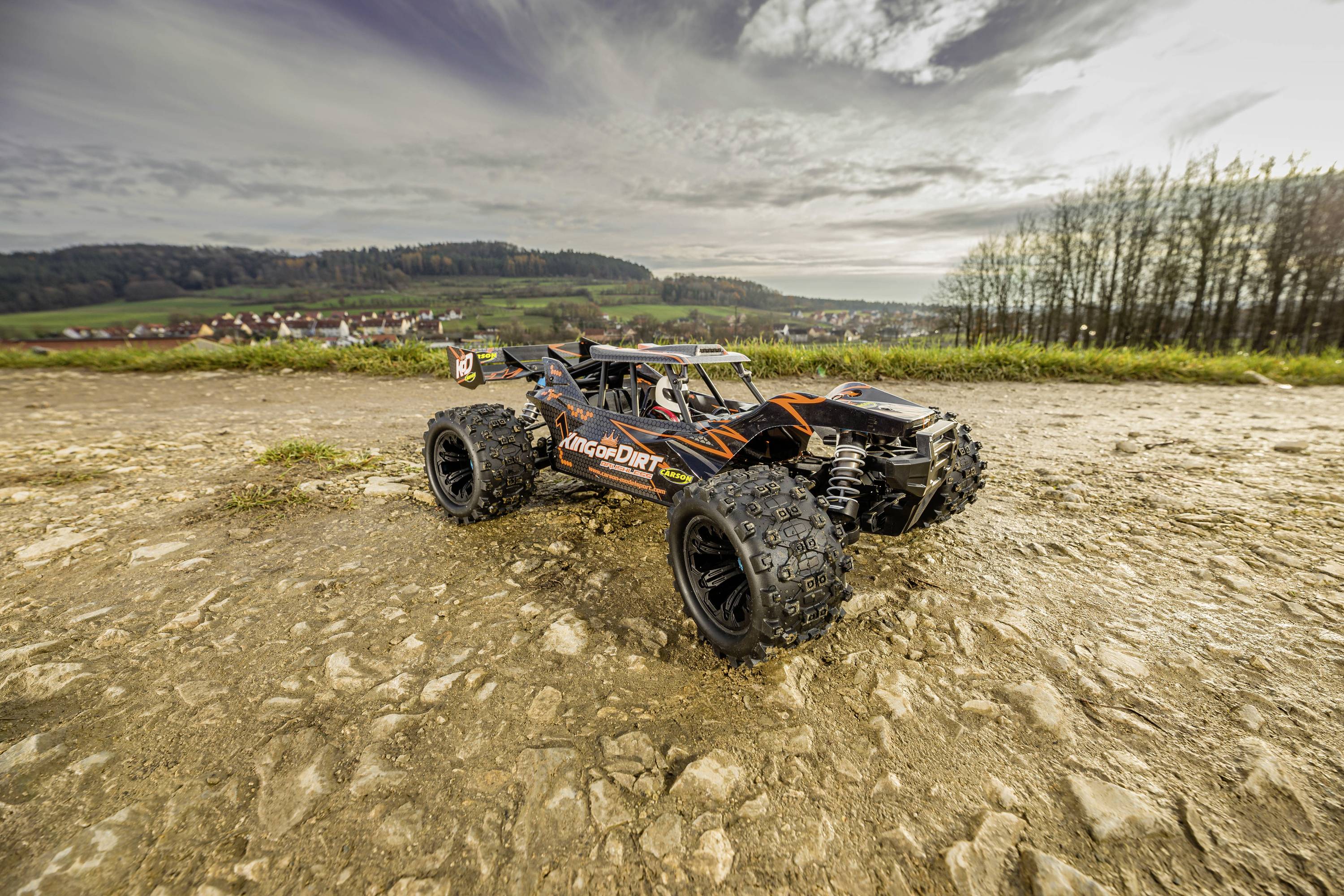 A remote-controlled car with robust tyres sits on a stony path against a landscape of hills and trees in the background.