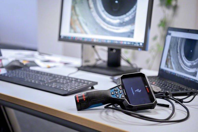 A portable endoscope device lies on a table next to a laptop and a monitor, both of which display enlarged pipe images.