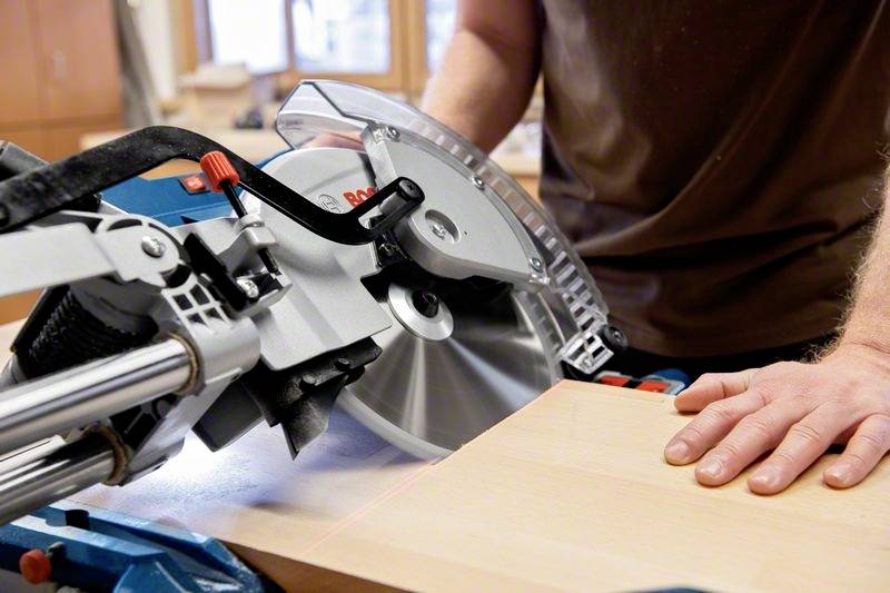 A person is cutting a wooden board with a mitre saw in a workshop. The blade is cutting precisely through the wood.