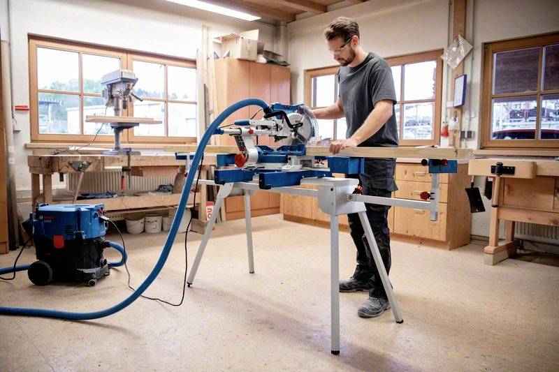 A man is cutting wood with a mitre saw in a workshop. A dust extraction system is connected to keep the workspace clean.