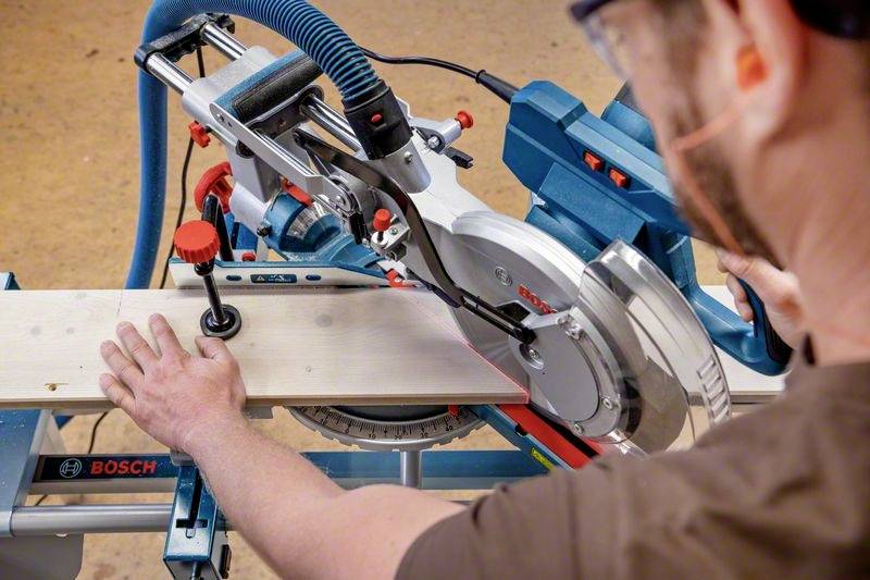 A person is cutting a wooden board with a mitre saw in a workshop. The focus is on the precise guiding of the saw.