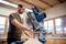 A man in a workshop is working with a blue mitre saw. Wooden shelves and large windows can be seen in the background.
