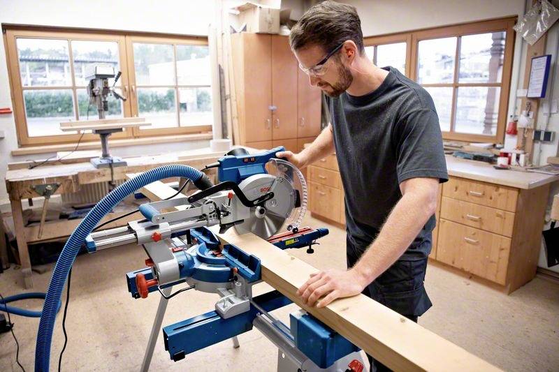 A man is working in a workshop with a blue mitre saw, cutting a piece of wood. Cupboards and windows are in the background.