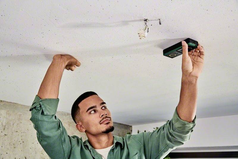 A man in a green shirt is measuring a ceiling with a hand tool. He appears focused and is working on a renovation.