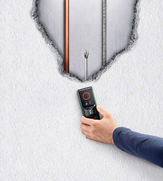 A hand is holding a digital detector against a wall that is broken through, revealing cables and pipes.