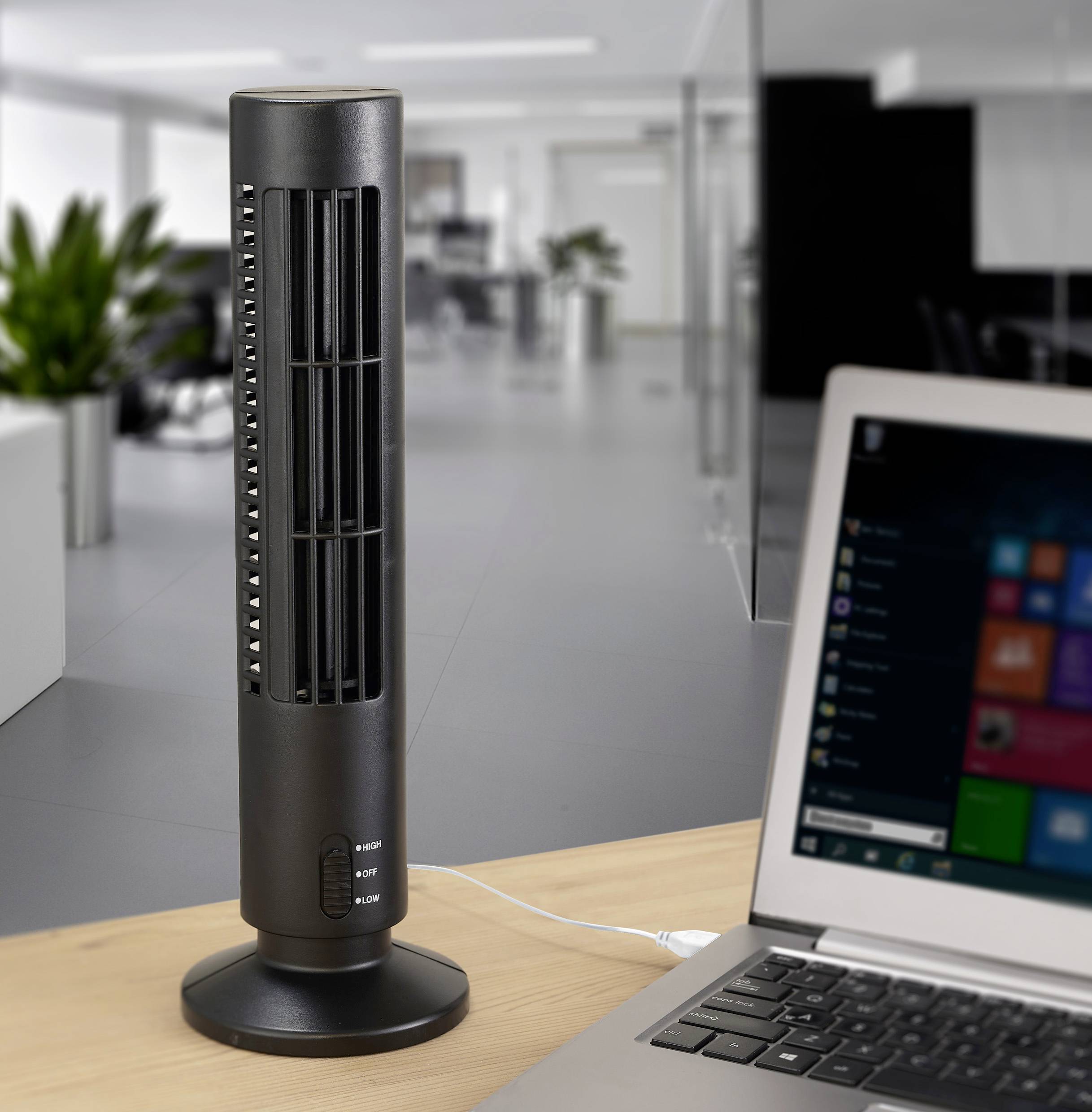 A black tower fan is switched on, standing on an office desk next to a laptop in a modern office with plants in the background.