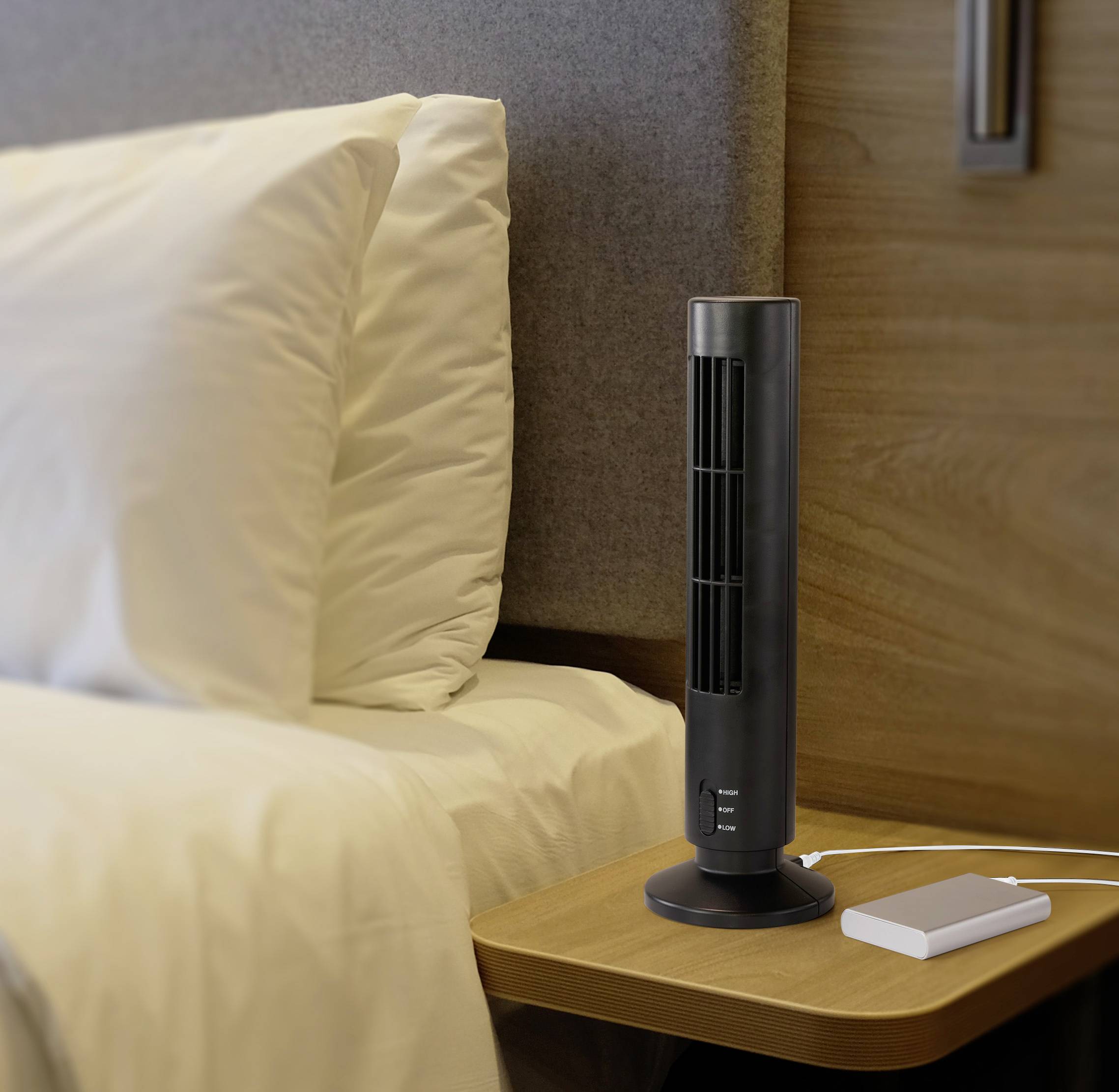 A small tower fan stands on a bedside table next to a bed fitted with white pillows.