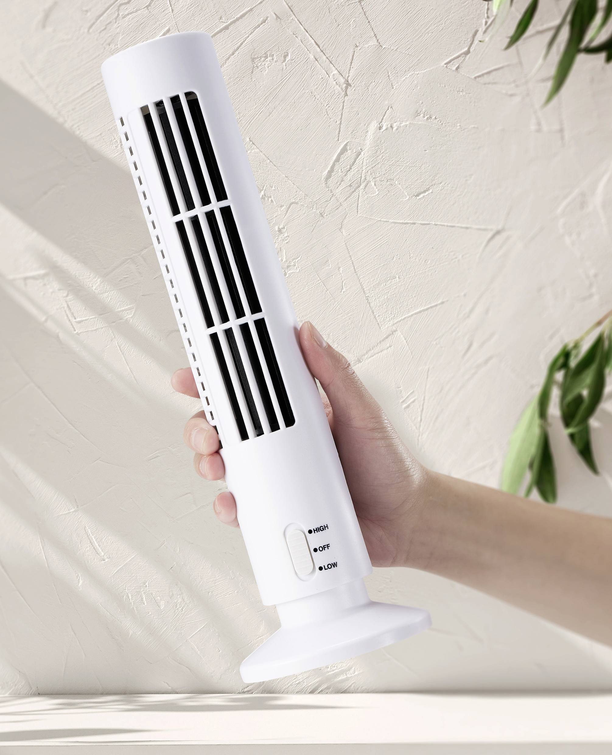 A hand is holding a slender, white desk fan in front of a textured, beige wall. The fan has three black grilles.