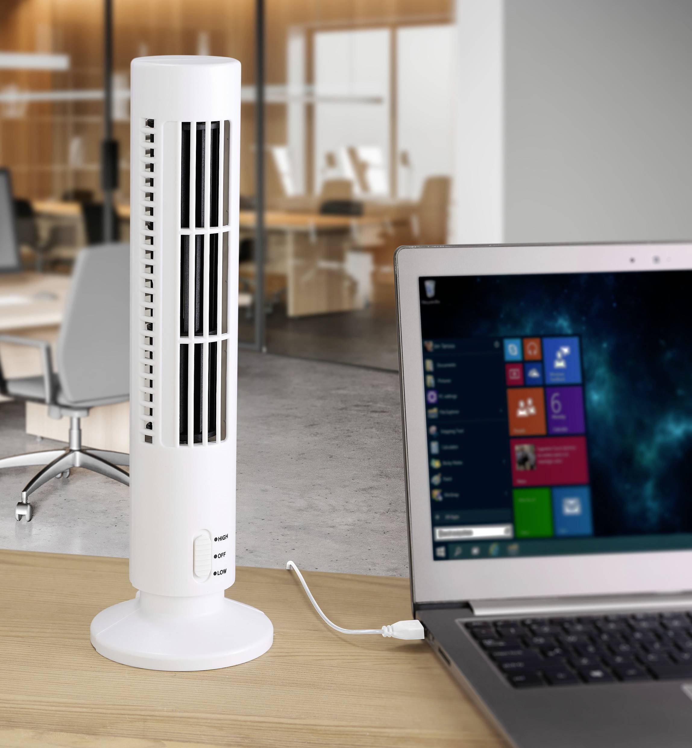 A white, slender fan sits on an office desk next to an open laptop. A modern office is visible in the background.
