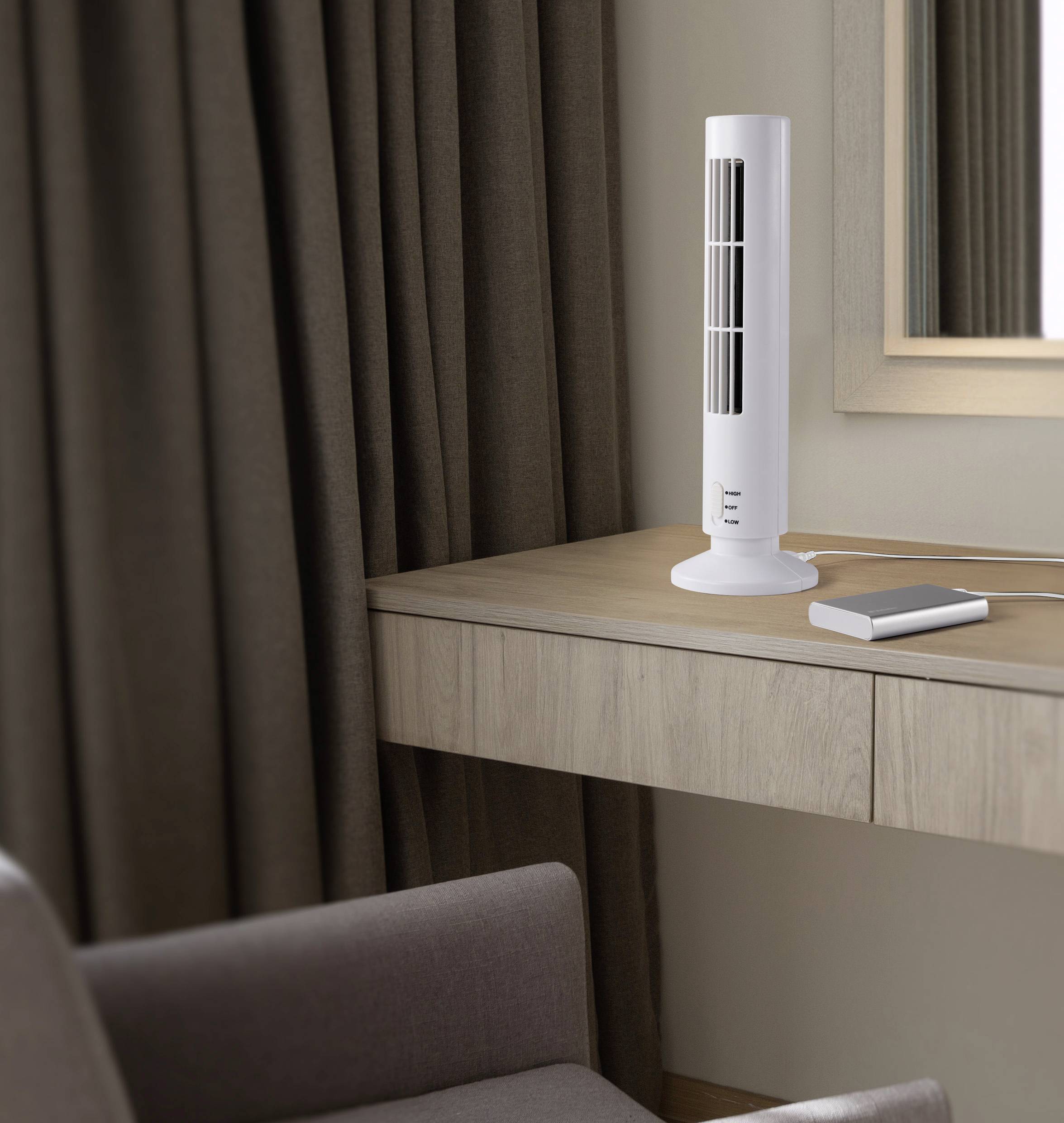 A desk fan sits on a light wooden table next to a notebook. Dark curtains and an armchair are in the background.