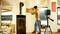 A man is aligning a wooden board on a living room wall using a laser device. In the background, there is a wood-burning stove and a patio.