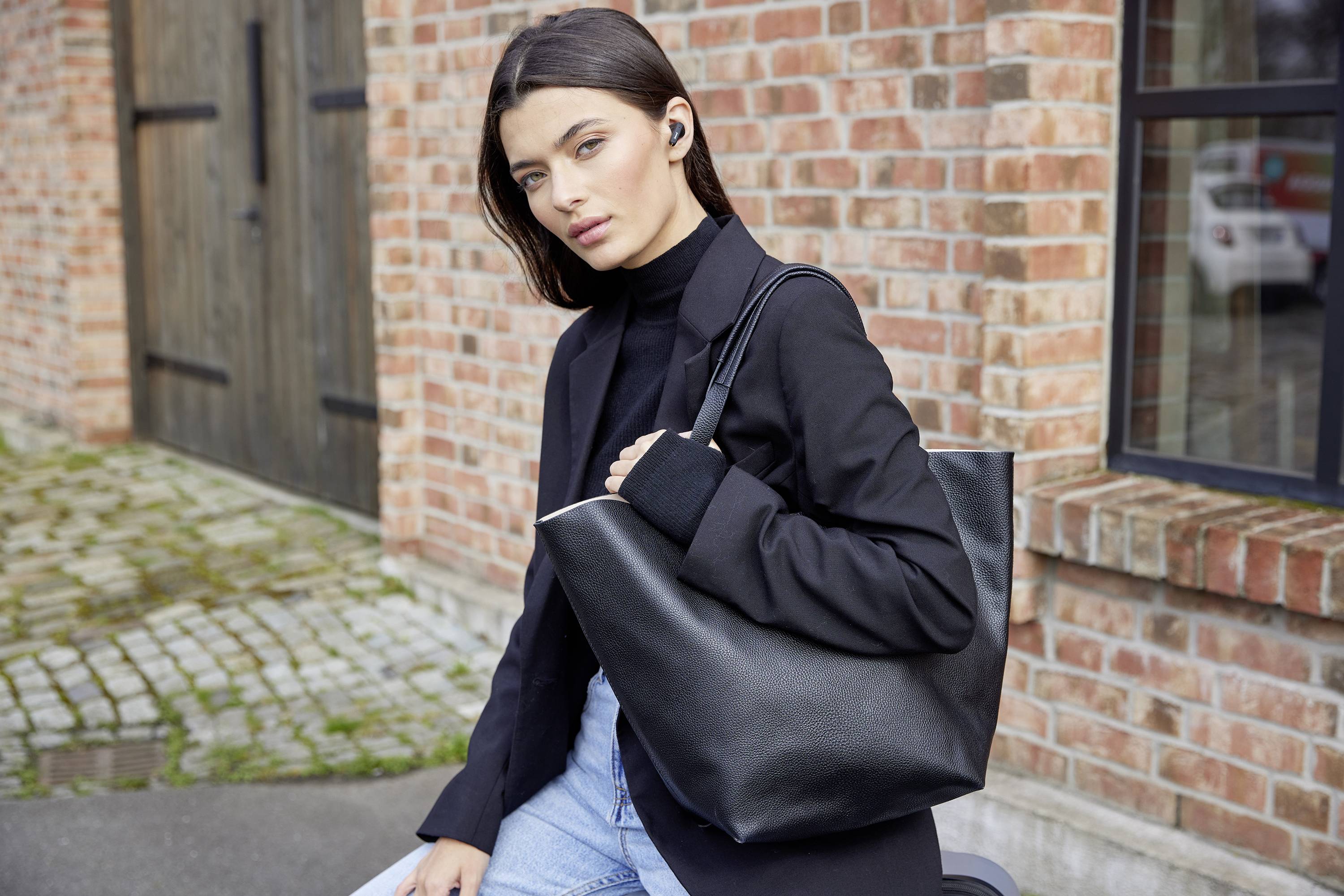 A woman dressed in black is sitting outside a brick building, carrying a large black bag on her shoulder.