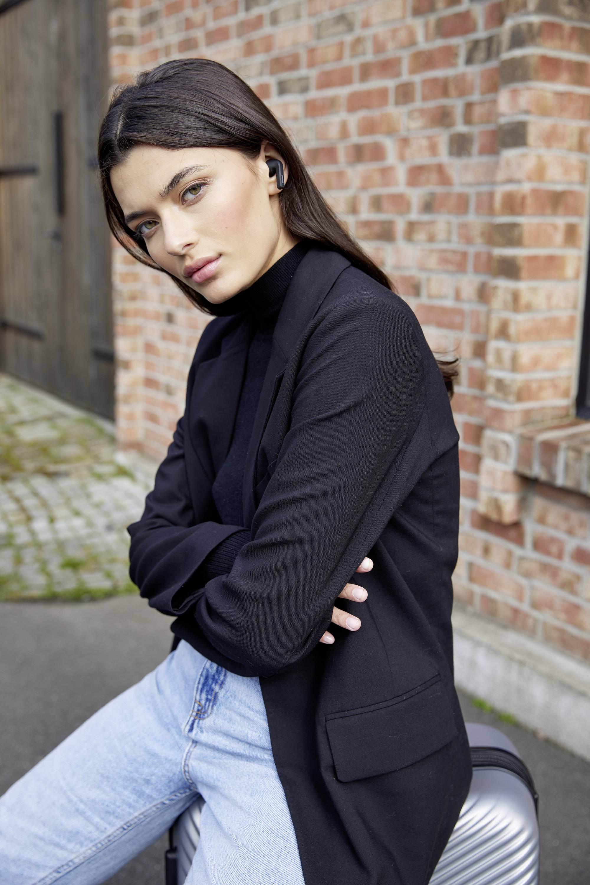 Woman in a black blazer and blue jeans leaning casually against a brick wall. She is wearing wireless earbuds and looks serious.