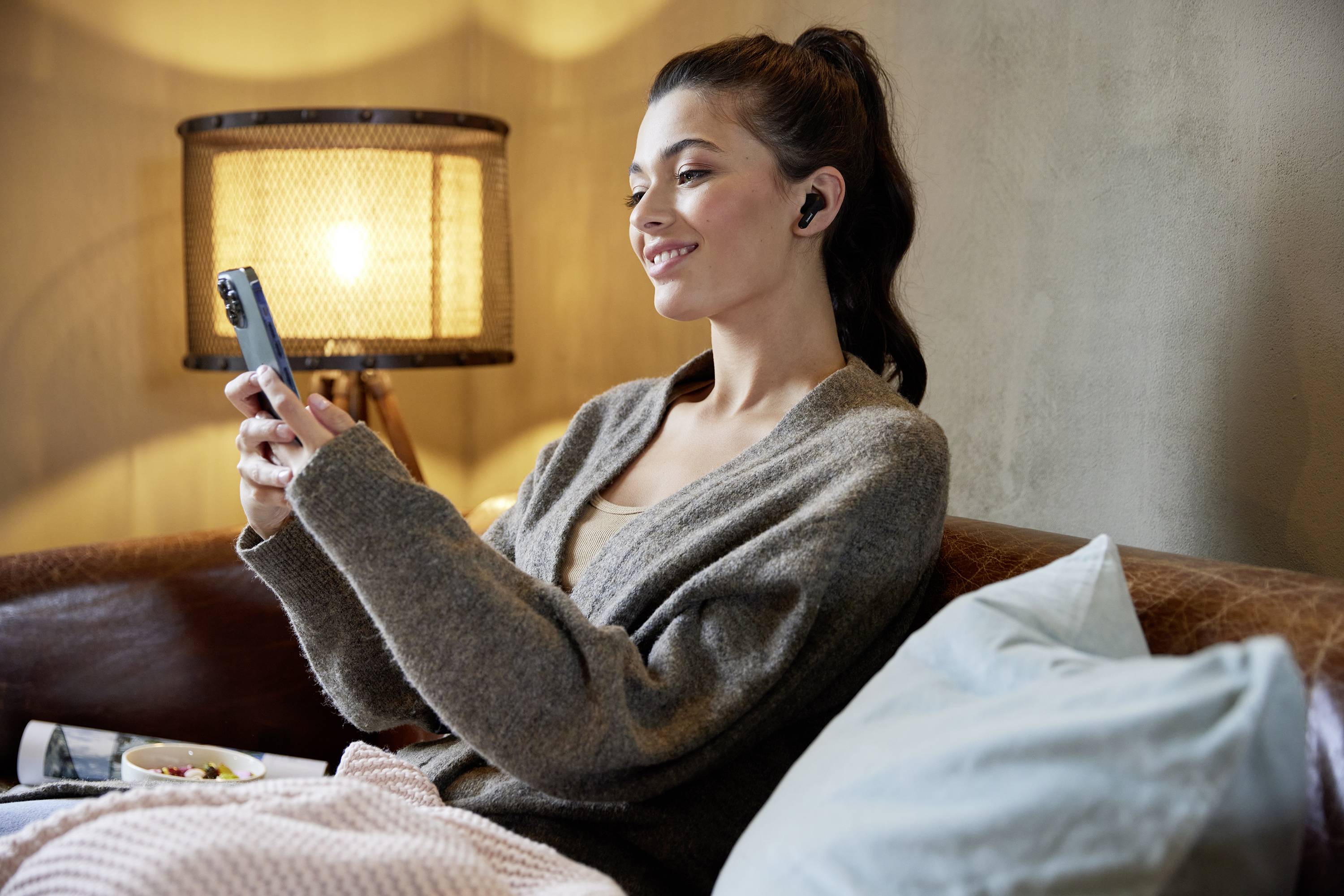 A smiling woman is sitting on a sofa, wearing wireless earphones and looking at her mobile phone. A lamp is illuminating the background.