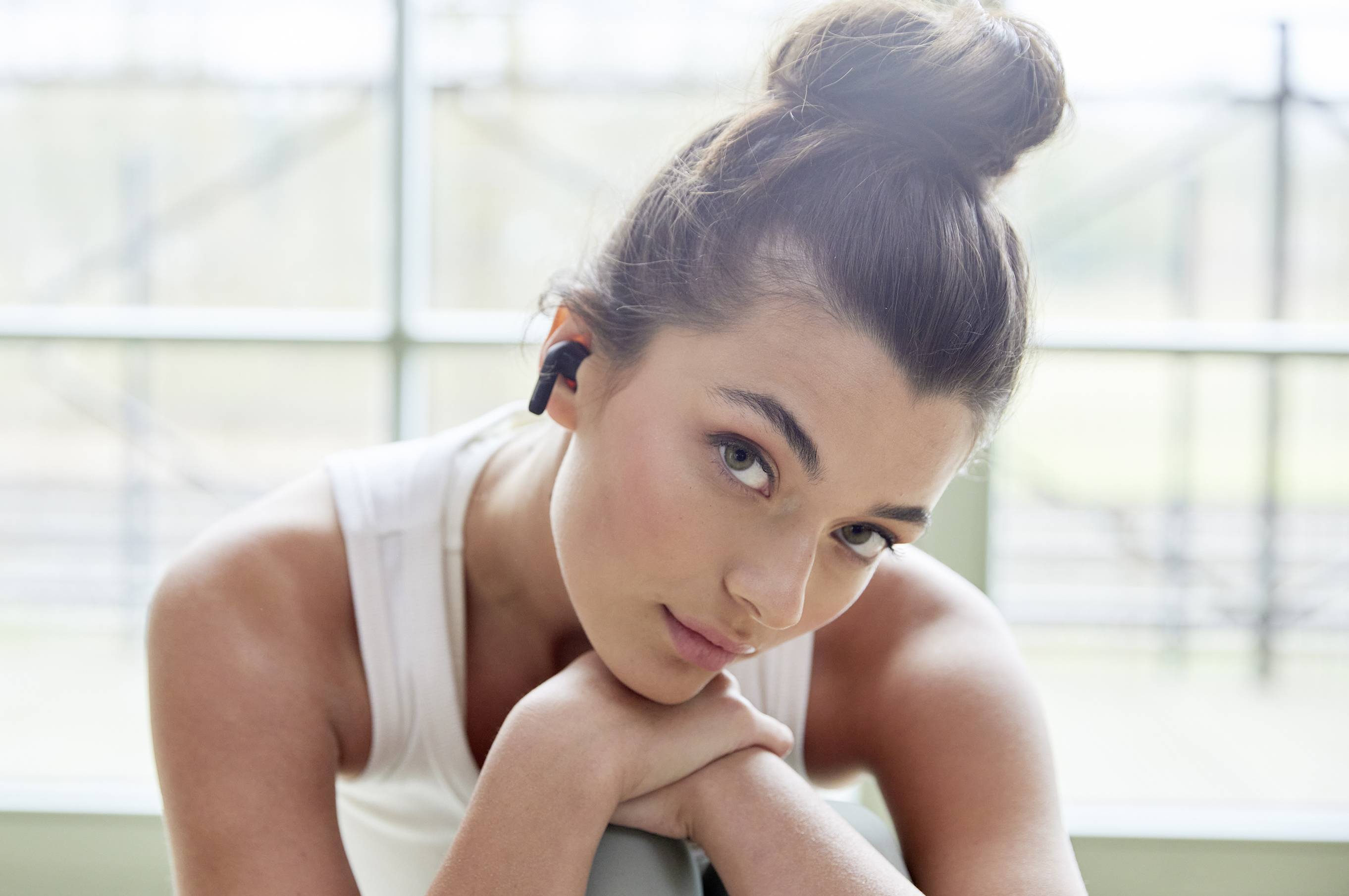 Young woman with dark hair wearing wireless earphones and resting her head on her hands. Background shows a window.