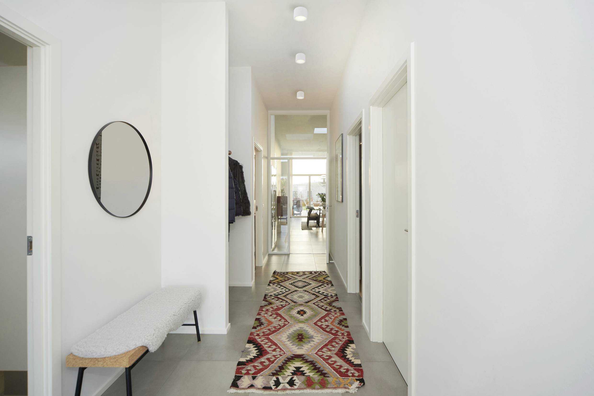 Long, bright hallway with a round mirror and a bench on the left side. A colourful carpet leads to a glass door at the back.