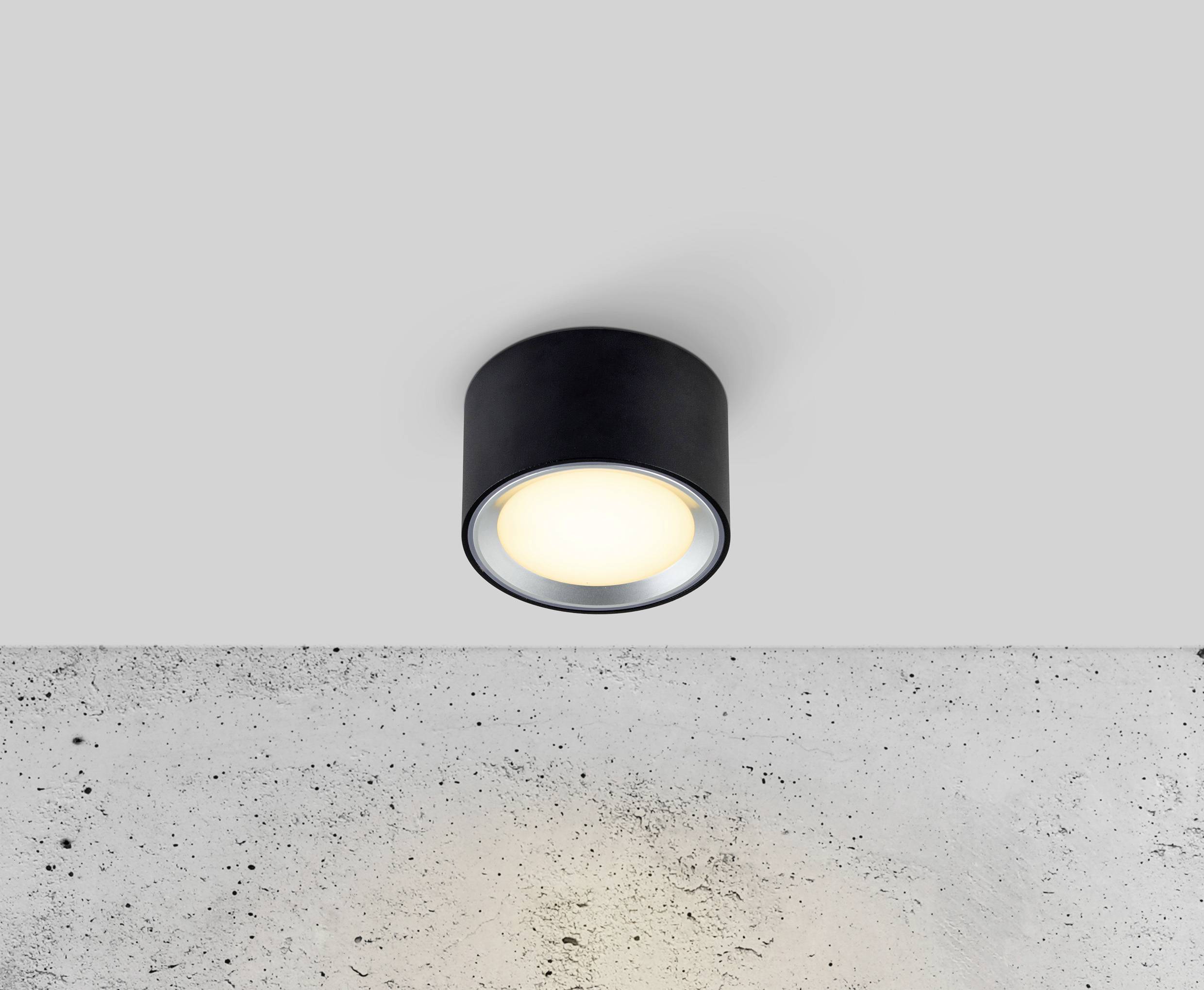 A round ceiling light is shining on a concrete surface. It has a simple, modern design and is emitting warm white light.