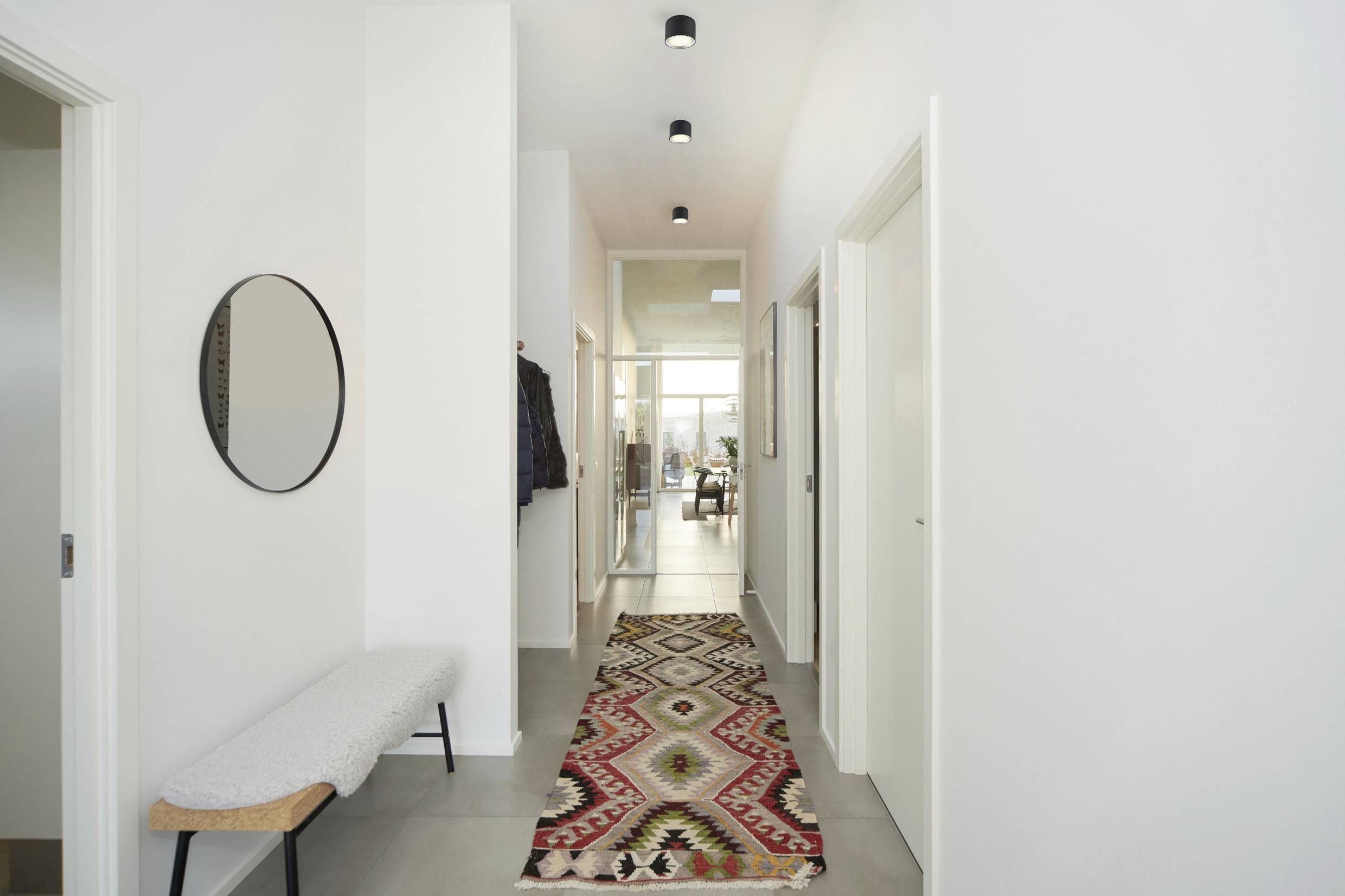 Long hallway with light decor. A round mirror hangs on the wall, with a bench underneath. A colourful runner covers the floor.