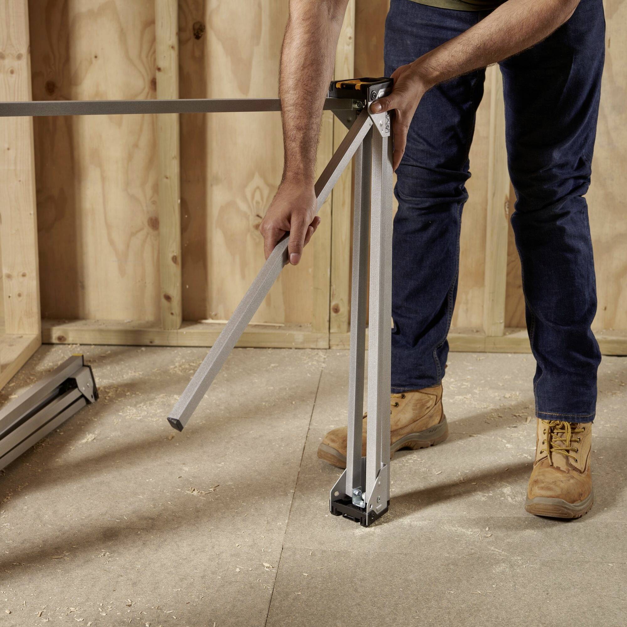 'A person adjusts the height of a table leg with a tool while standing in an unfinished room with wooden panelling.'