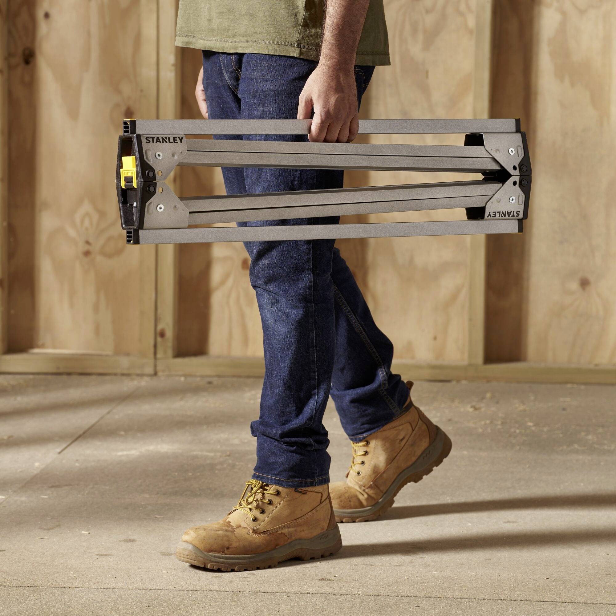 A person in workwear is carrying a folded sawhorse in a workshop environment. Wooden walls in the background.