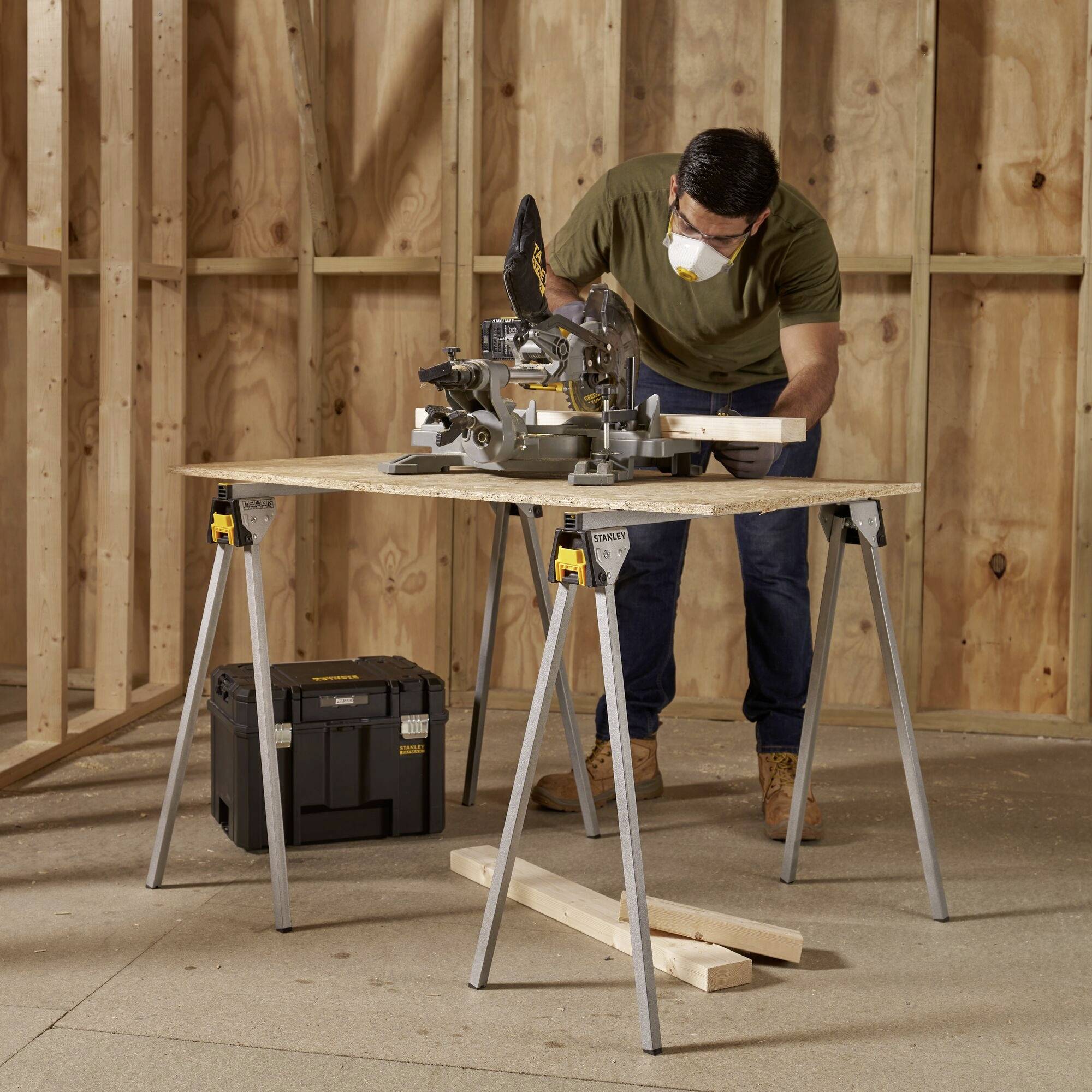 A person is cutting wood with a mitre saw in a workshop. They are wearing a safety mask. Wooden walls can be seen in the background.