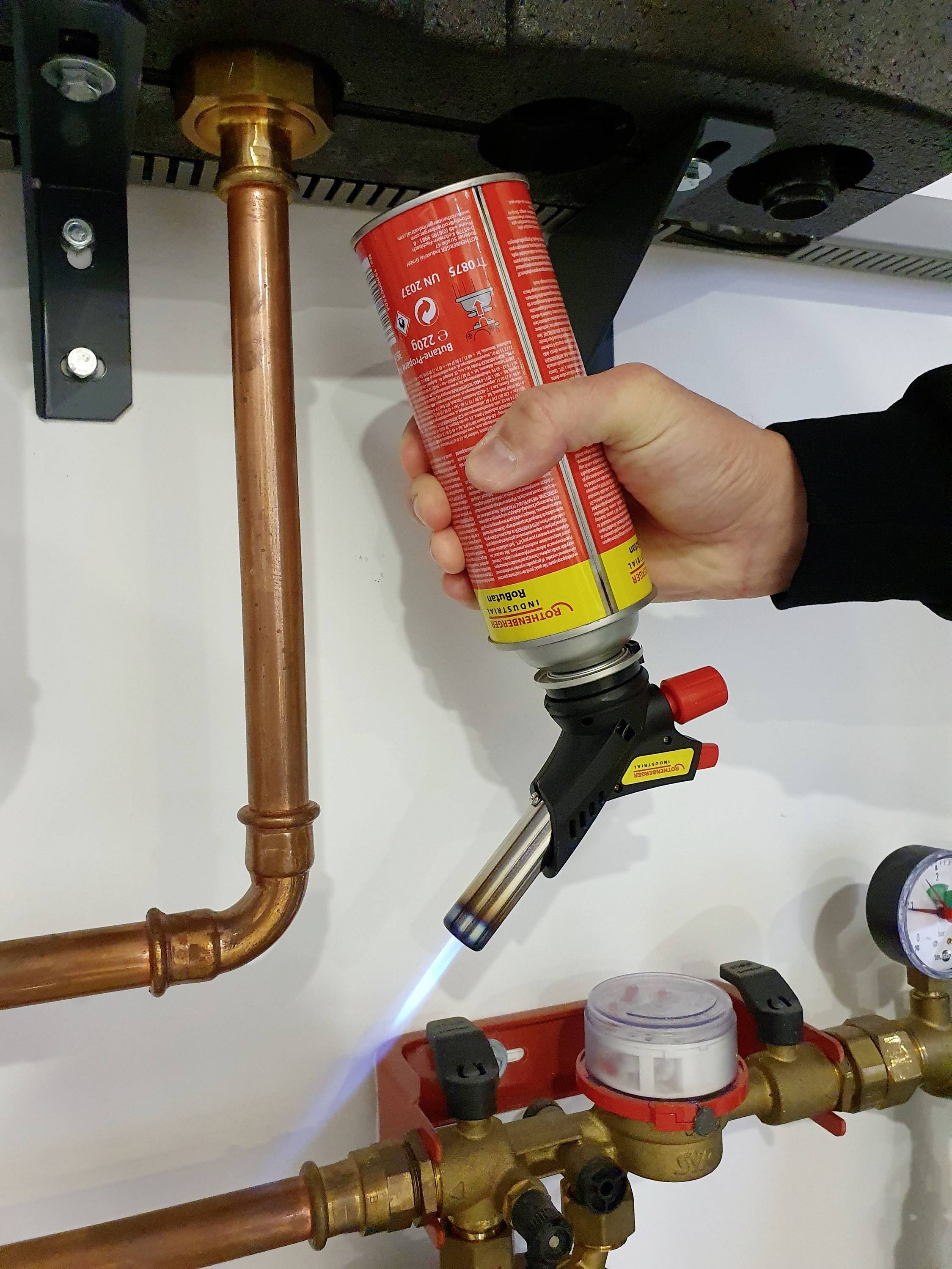 A hand holds a blowtorch, heating a copper pipe in a heating system. A pressure gauge is visible in the background.