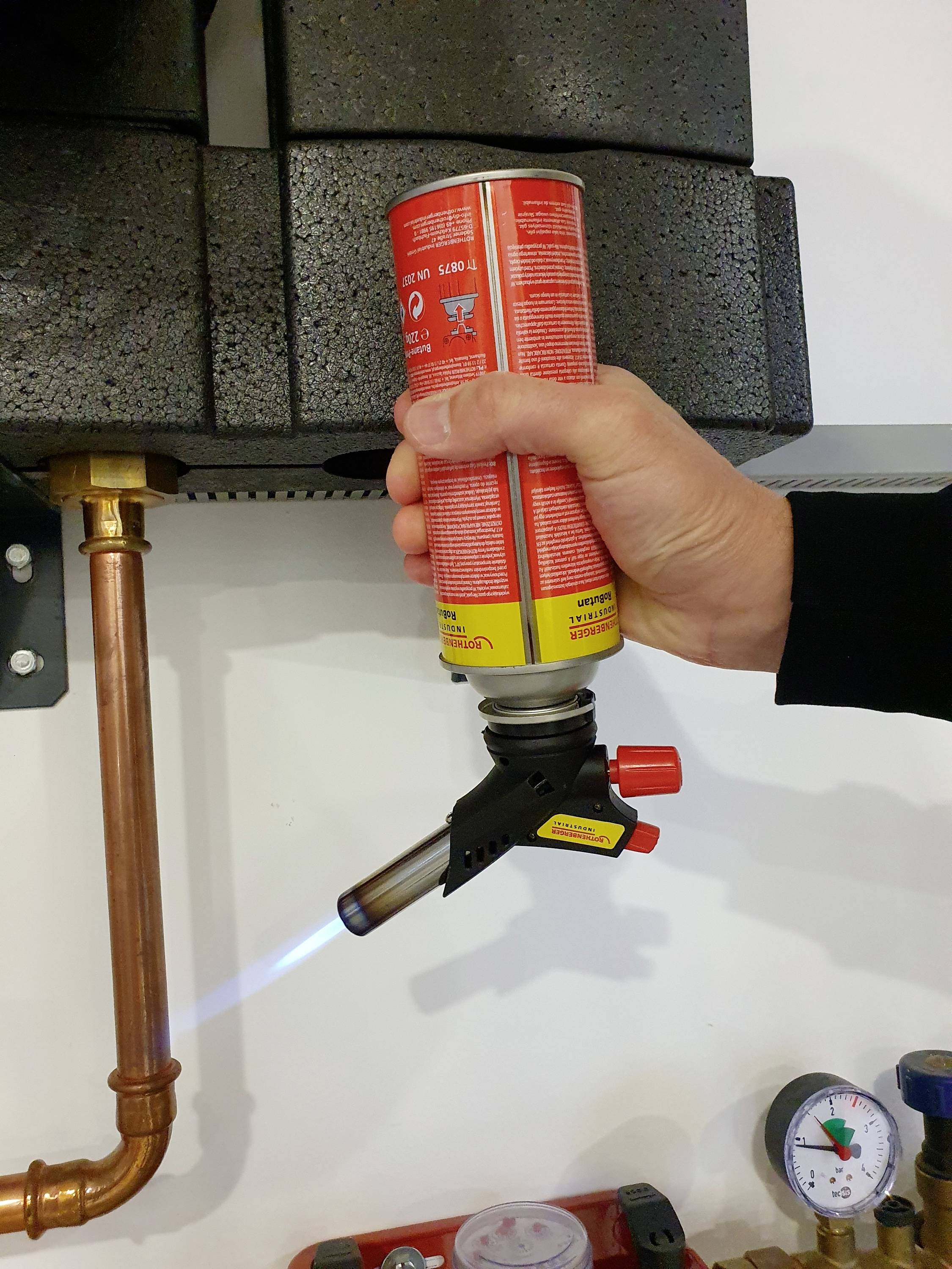 A hand holds a blowtorch with a blue flame against a copper pipe. Next to the pipe is a pressure gauge.
