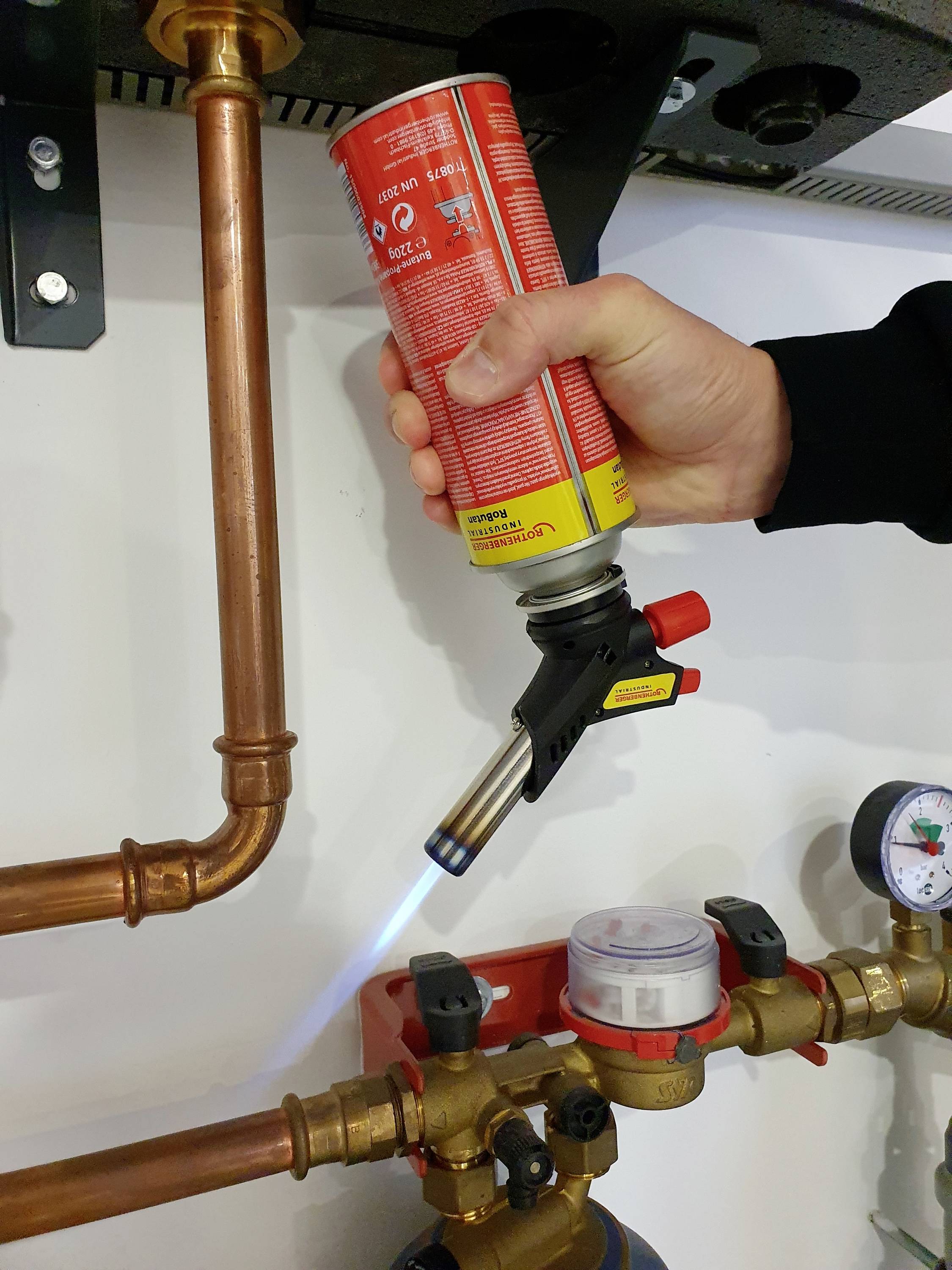 A hand holds a blowtorch canister, directing a blue flame towards a pipe valve. Copper pipes and pressure gauges are visible.